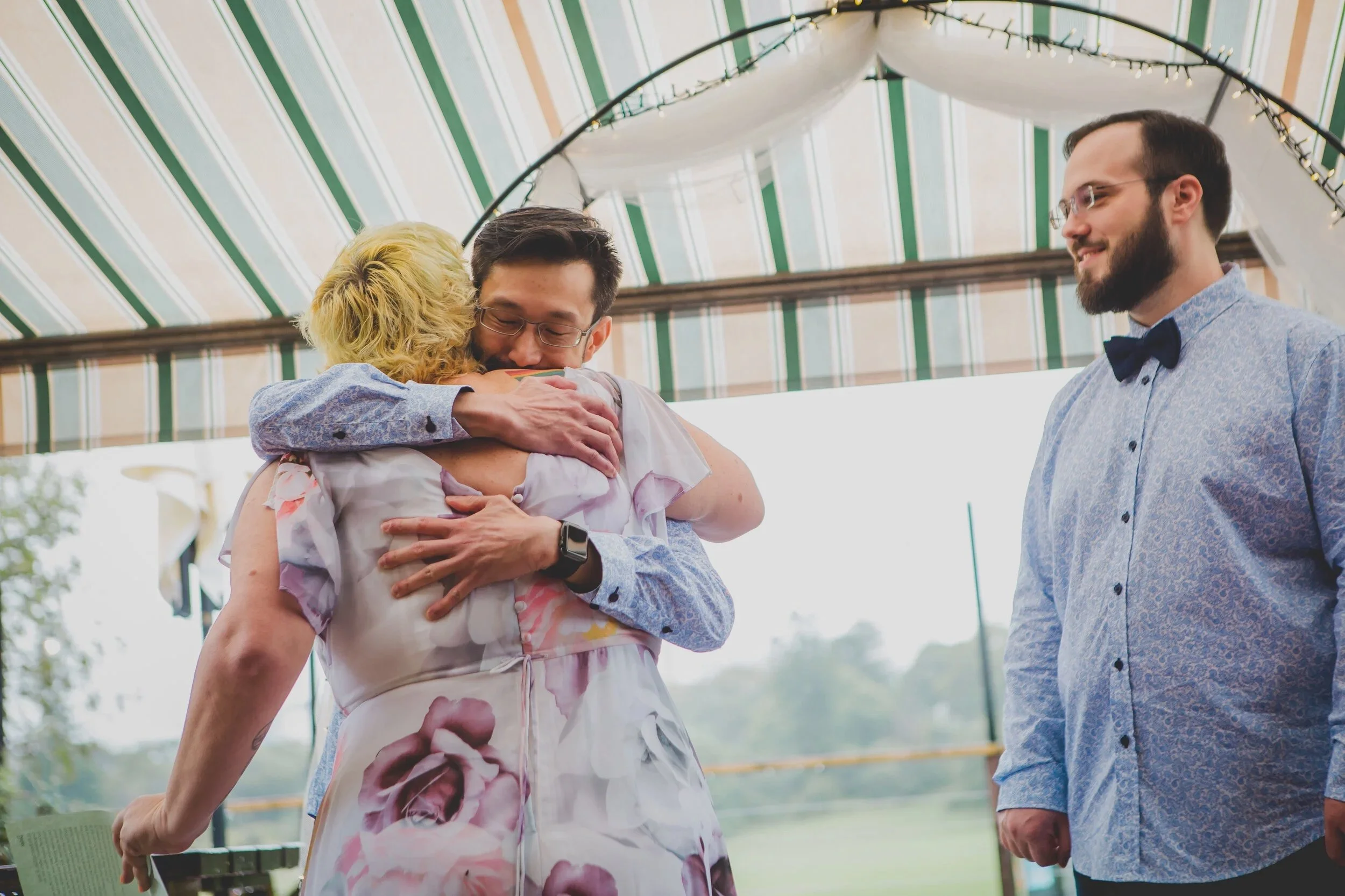 Two people hugging, with a third person standing and smiling nearby, at an indoor event with a striped awning and string lights overhead.