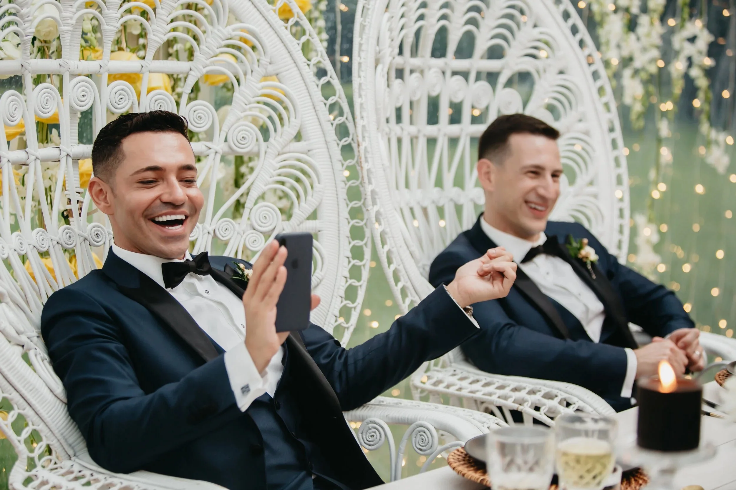 Two men dressed in tuxedos at a wedding reception, sitting on white wicker chairs with intricate designs, surrounded by fairy lights and flowers, smiling and enjoying the moment.