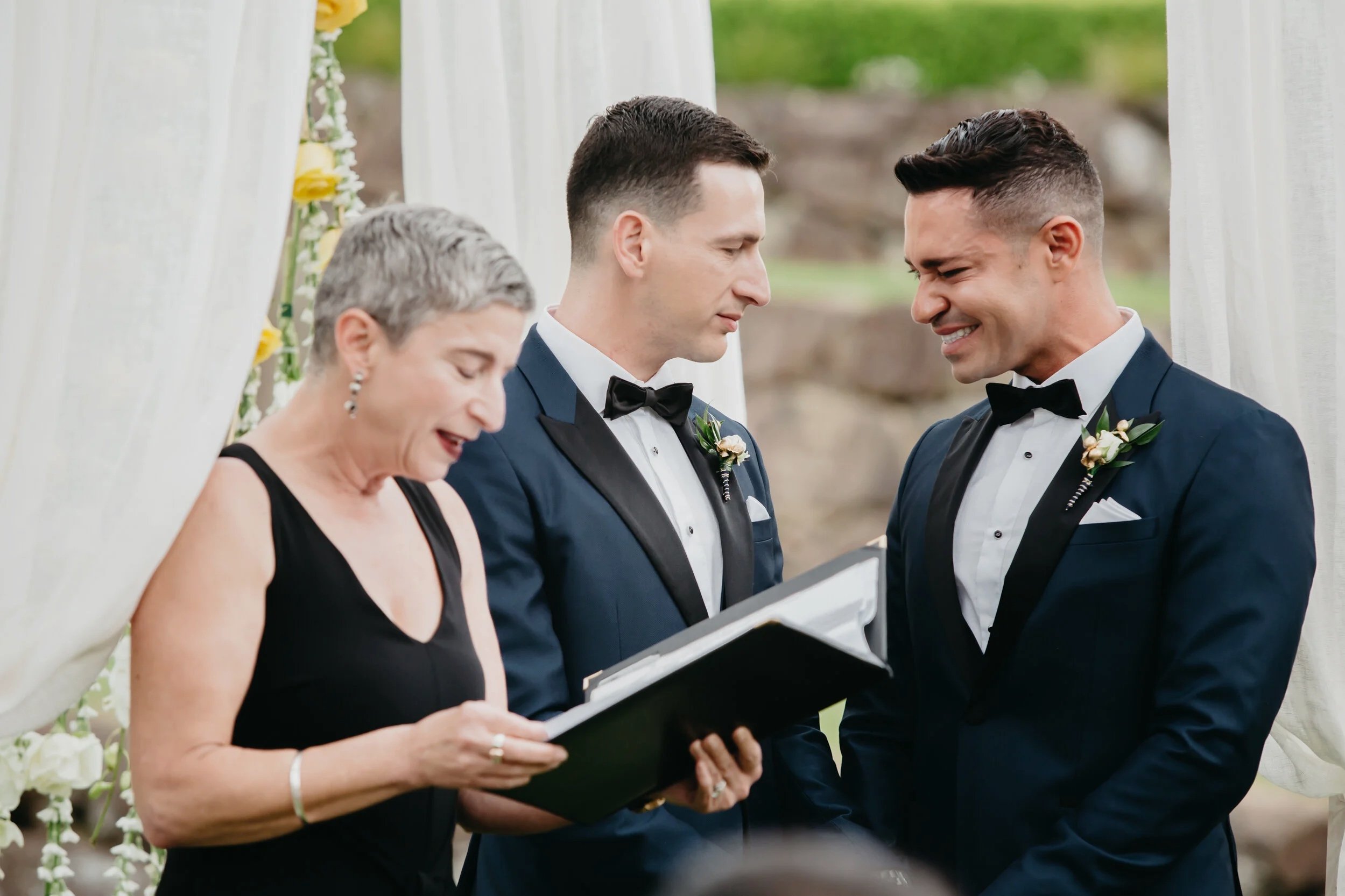 A same-sex couple getting married outdoors, reading vows with an officiant, during a wedding ceremony under a decorated archway.