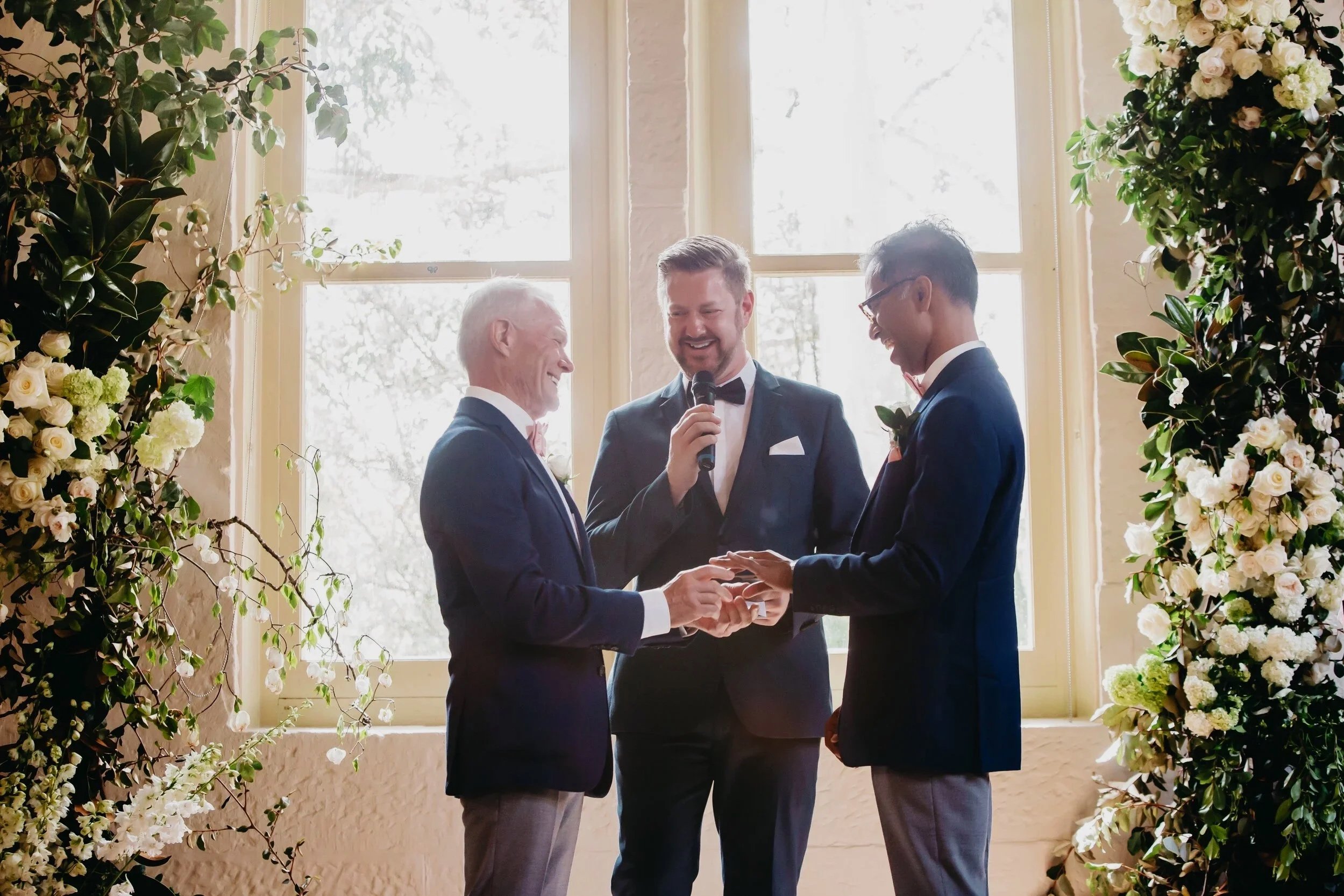 A same-sex couple is holding hands during their wedding ceremony, exchanging vows in front of officiant and surrounded by floral arrangements.