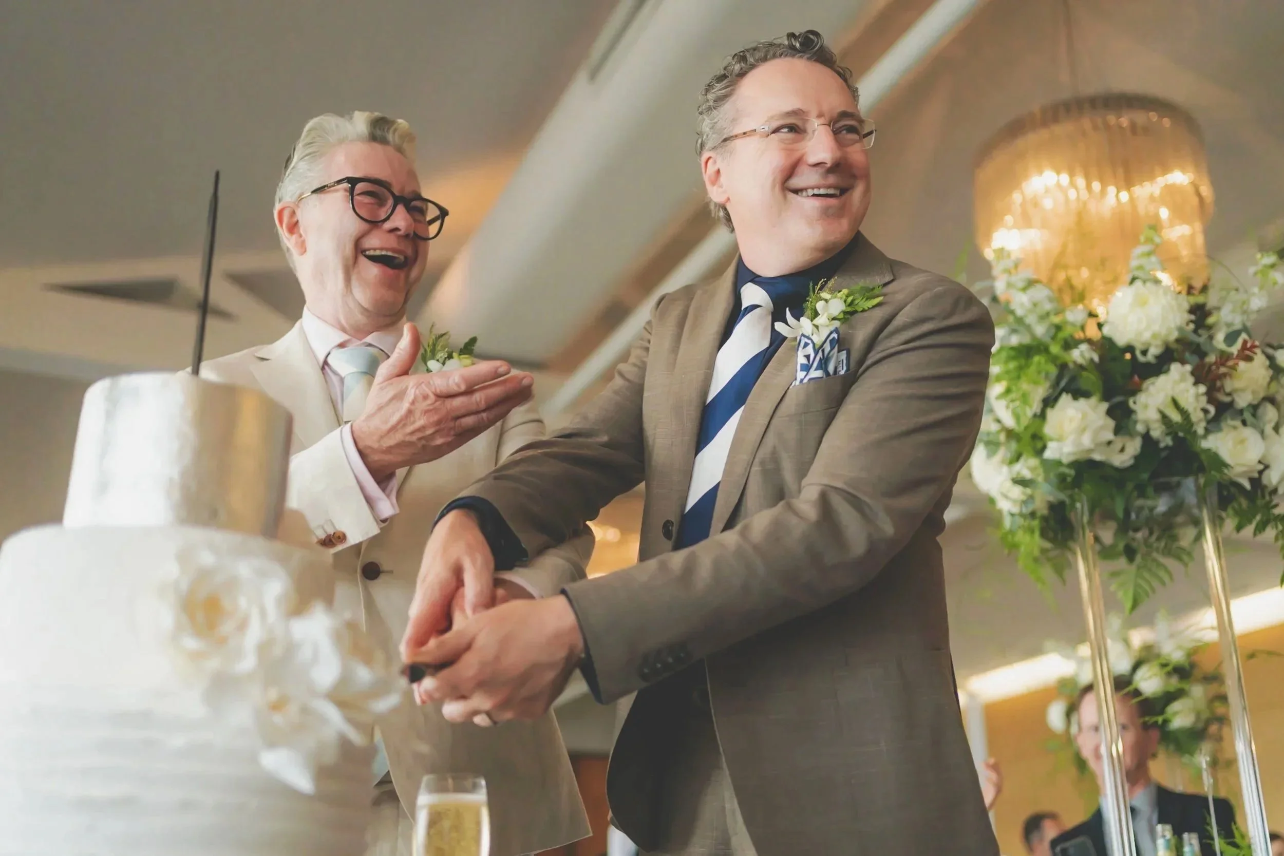 Two men in suits at a wedding celebration, one is laughing and the other is smiling, holding hands near a wedding cake, with floral arrangements in the background.
