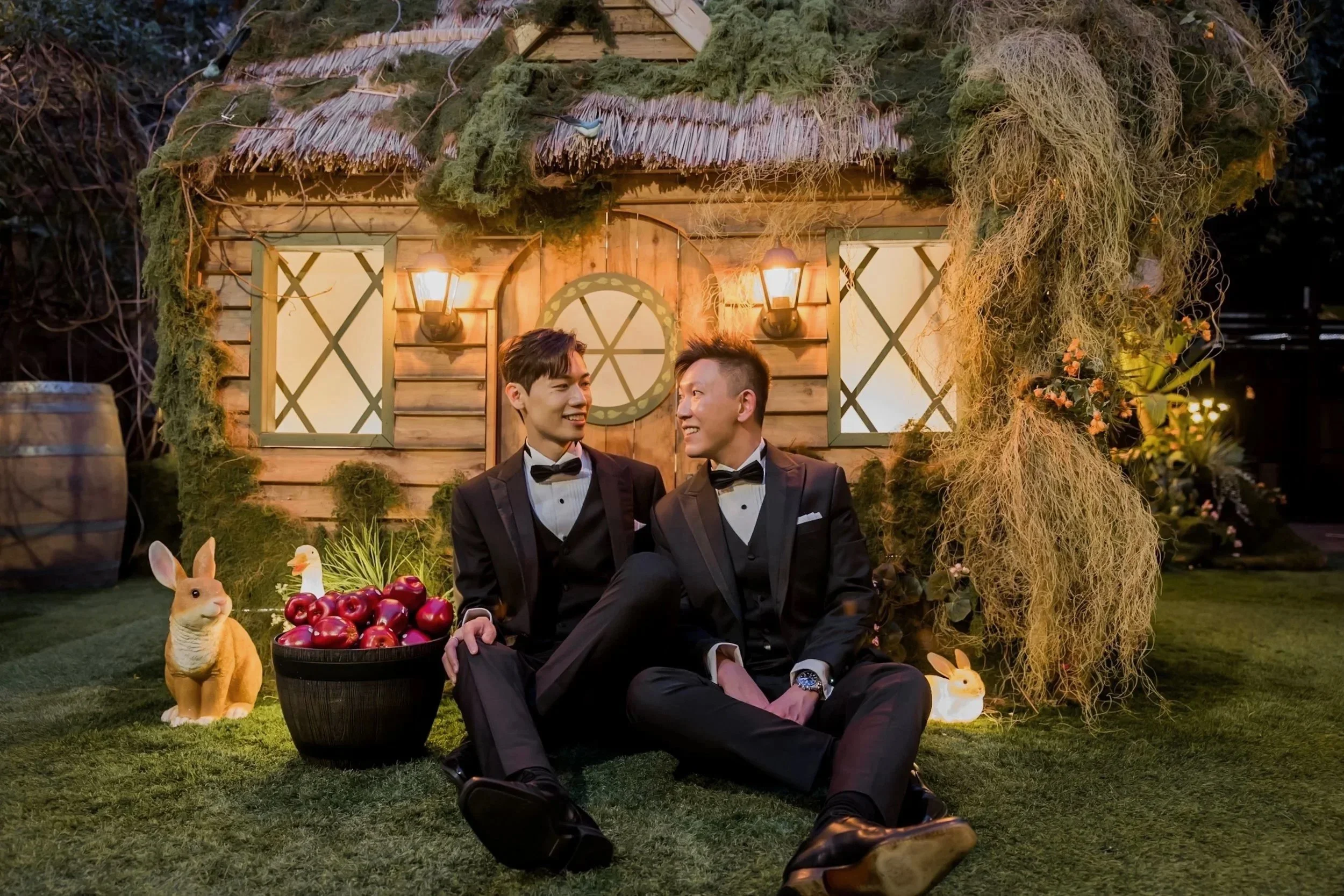 Two men in tuxedos sitting on grass in front of a whimsical wooden house with lantern lights, surrounded by bunny and duck decorations, and a basket of red apples.