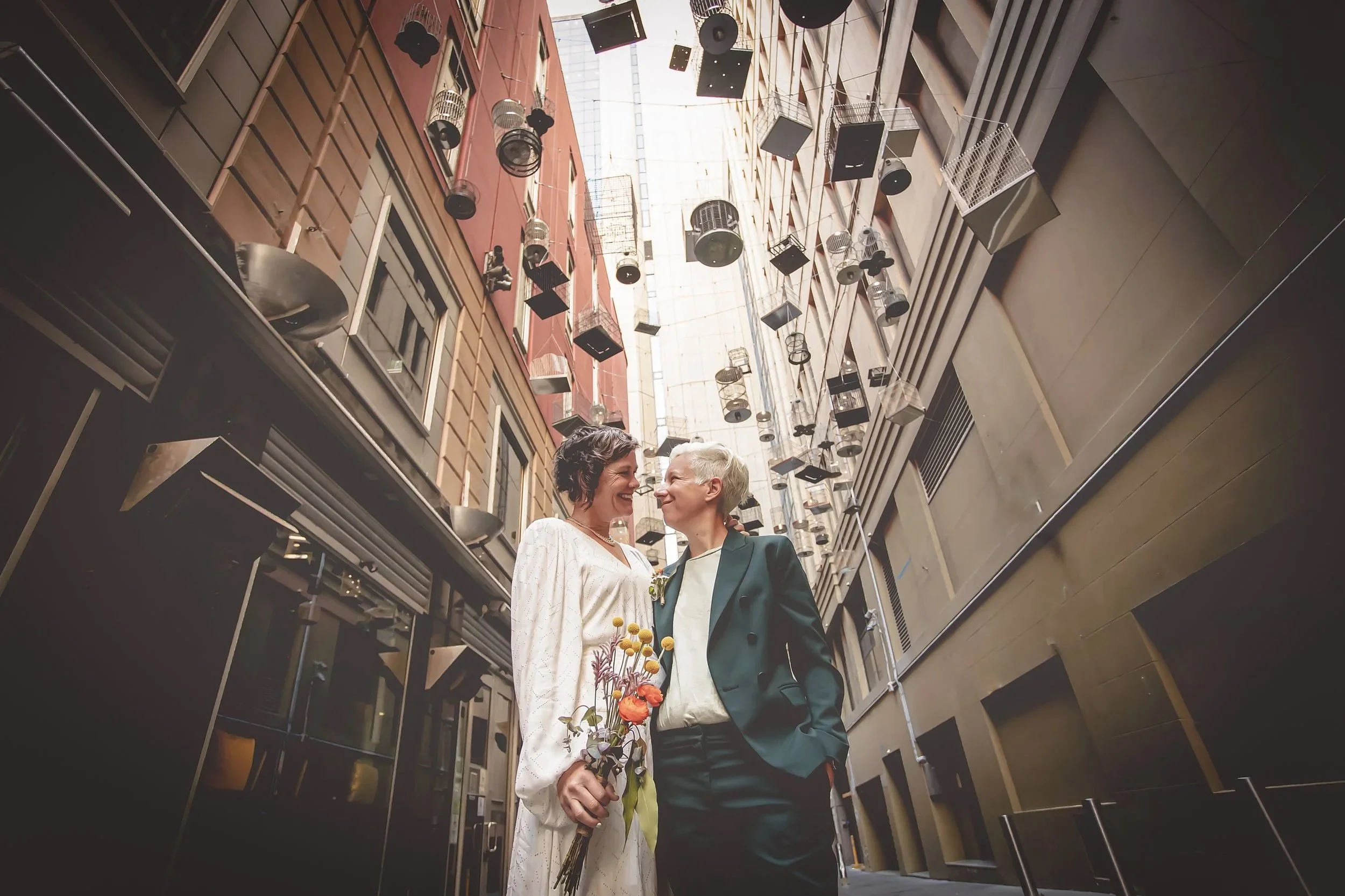 Two women stand close together in an urban alleyway, smiling at each other. One woman is holding a bouquet of flowers. Above them, birdcages and speakers are hanging from the buildings' facades.