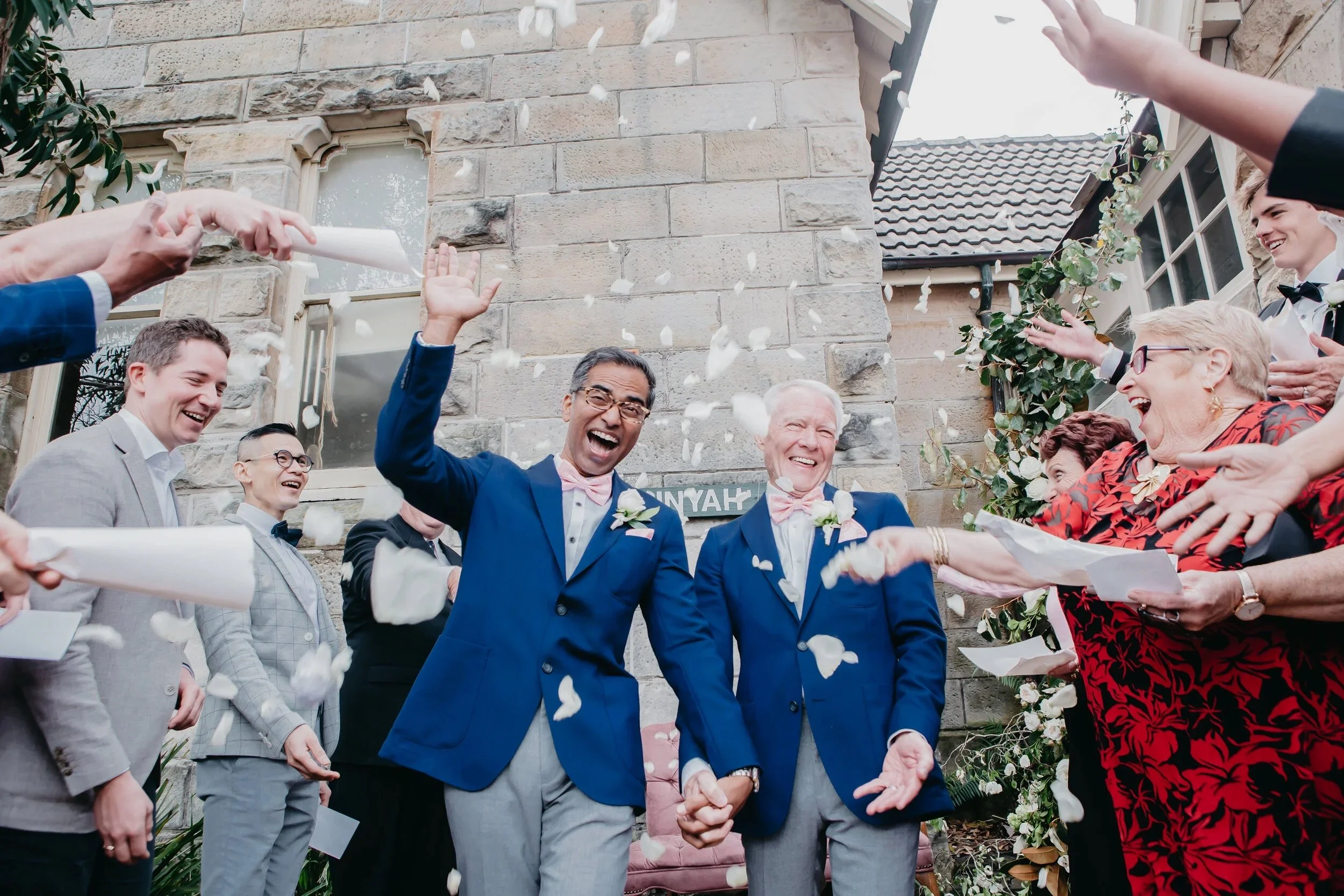Two men in blue suits and pink bowties holding hands and laughing as they are showered with white flower petals outside a stone building during a wedding celebration.