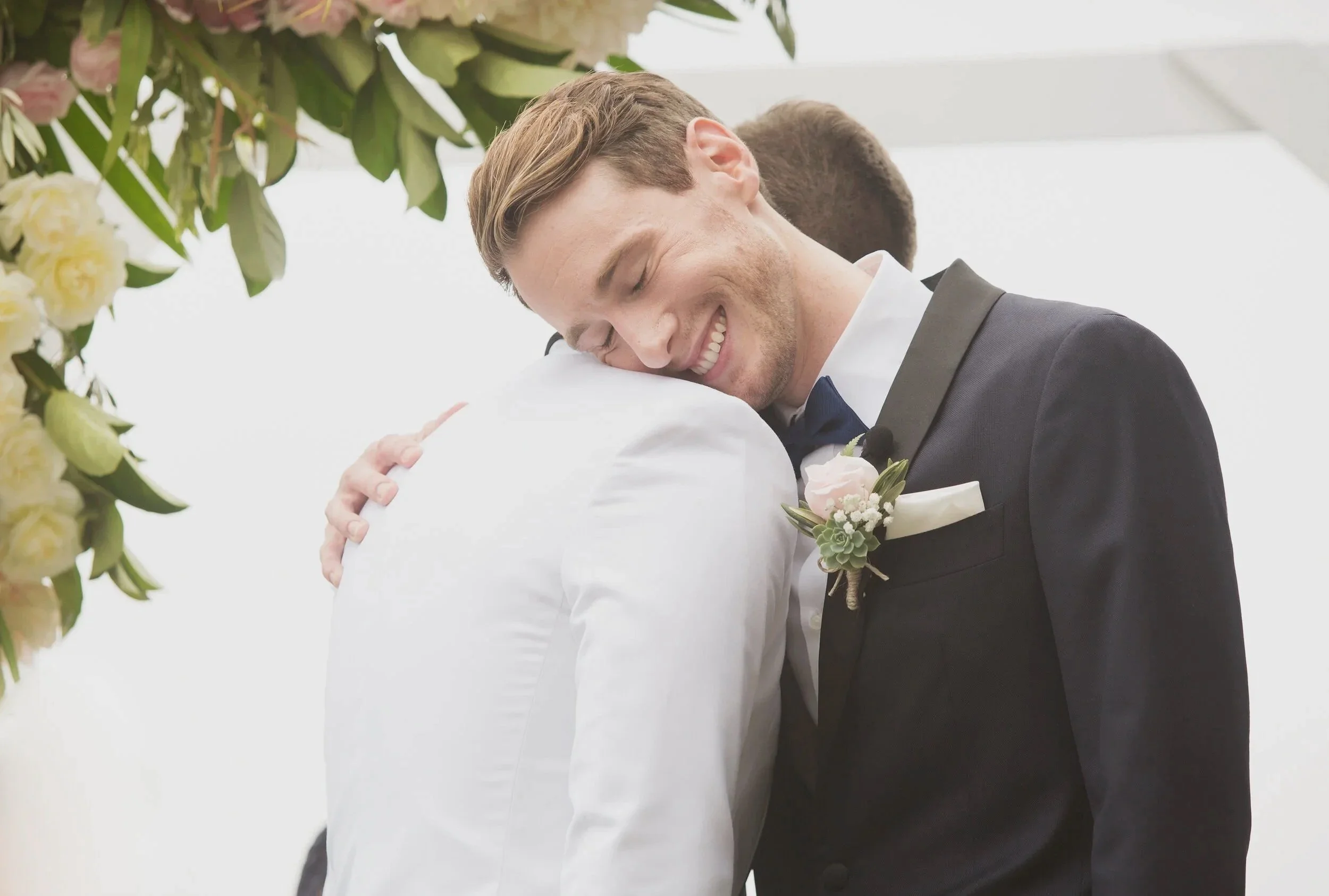 A man in a dark suit, white shirt, and dark tie is happily hugging another man in a white shirt at a wedding, with floral decorations in the background.