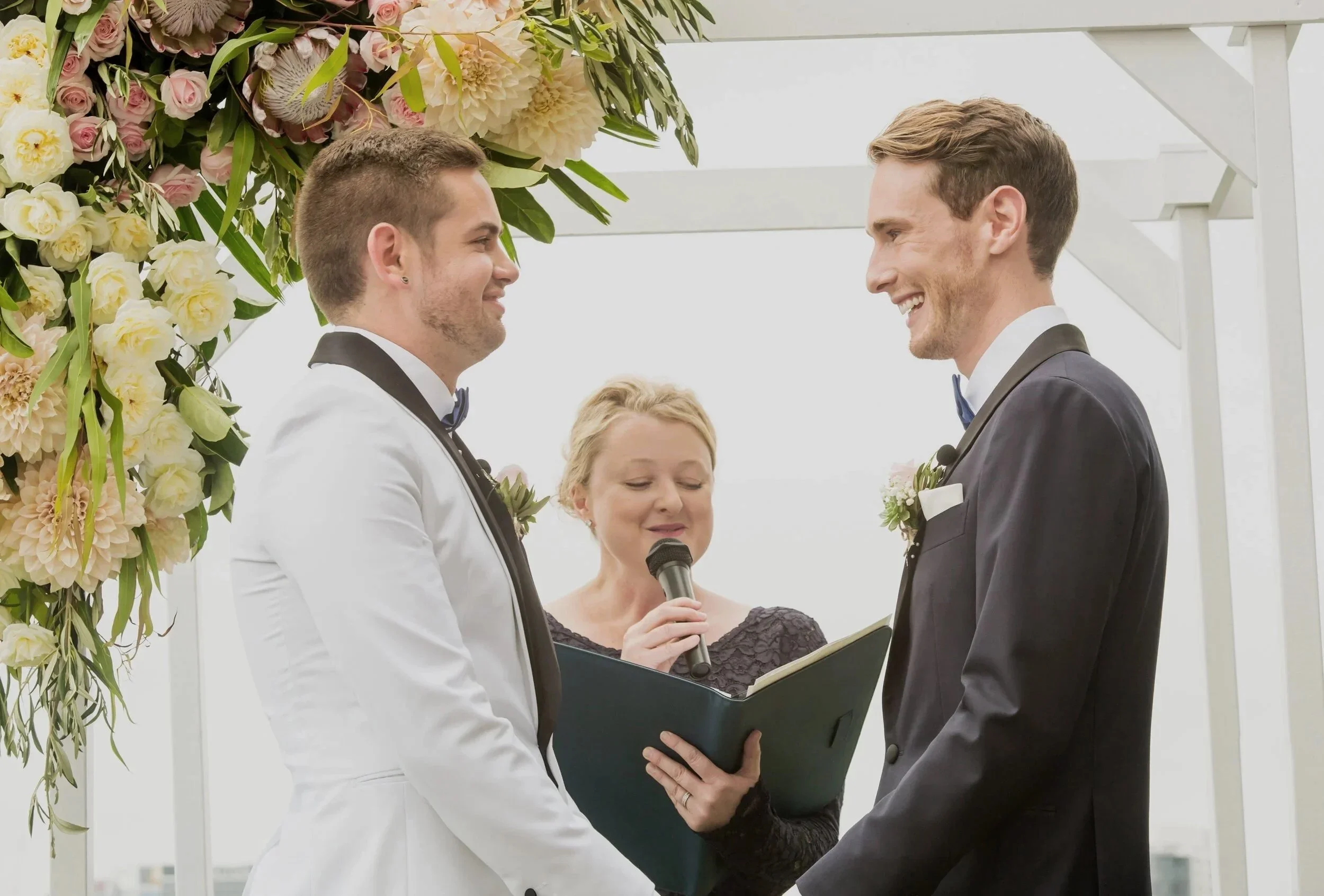 Two men in tuxedos holding hands during a wedding ceremony, with a woman officiant reading from a book, under a floral arch with white and pink flowers.