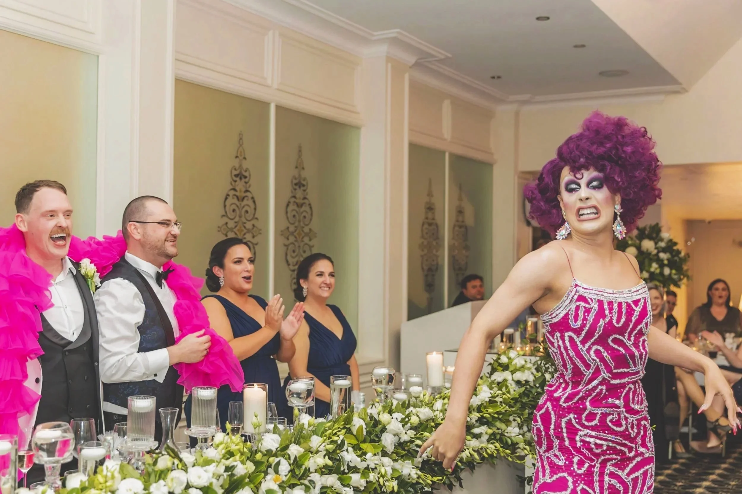 A drag performer in a pink and silver dress with the words 'GAY' and 'TOO' on it, performing at a wedding reception, with guests clapping and smiling in the background.