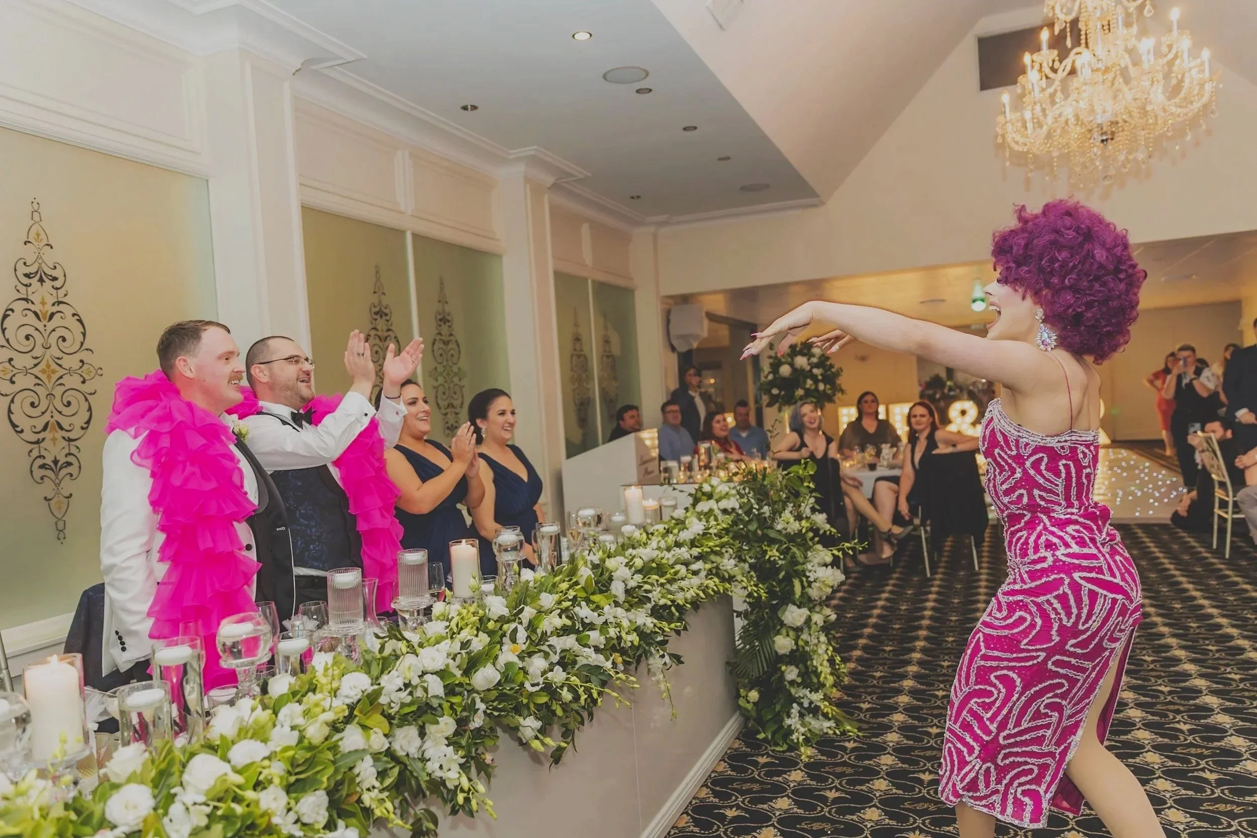 A lively celebration featuring a performer with a curly purple wig and pink dress entertaining guests sitting at a decorated table with candles and floral arrangements in an elegant venue.