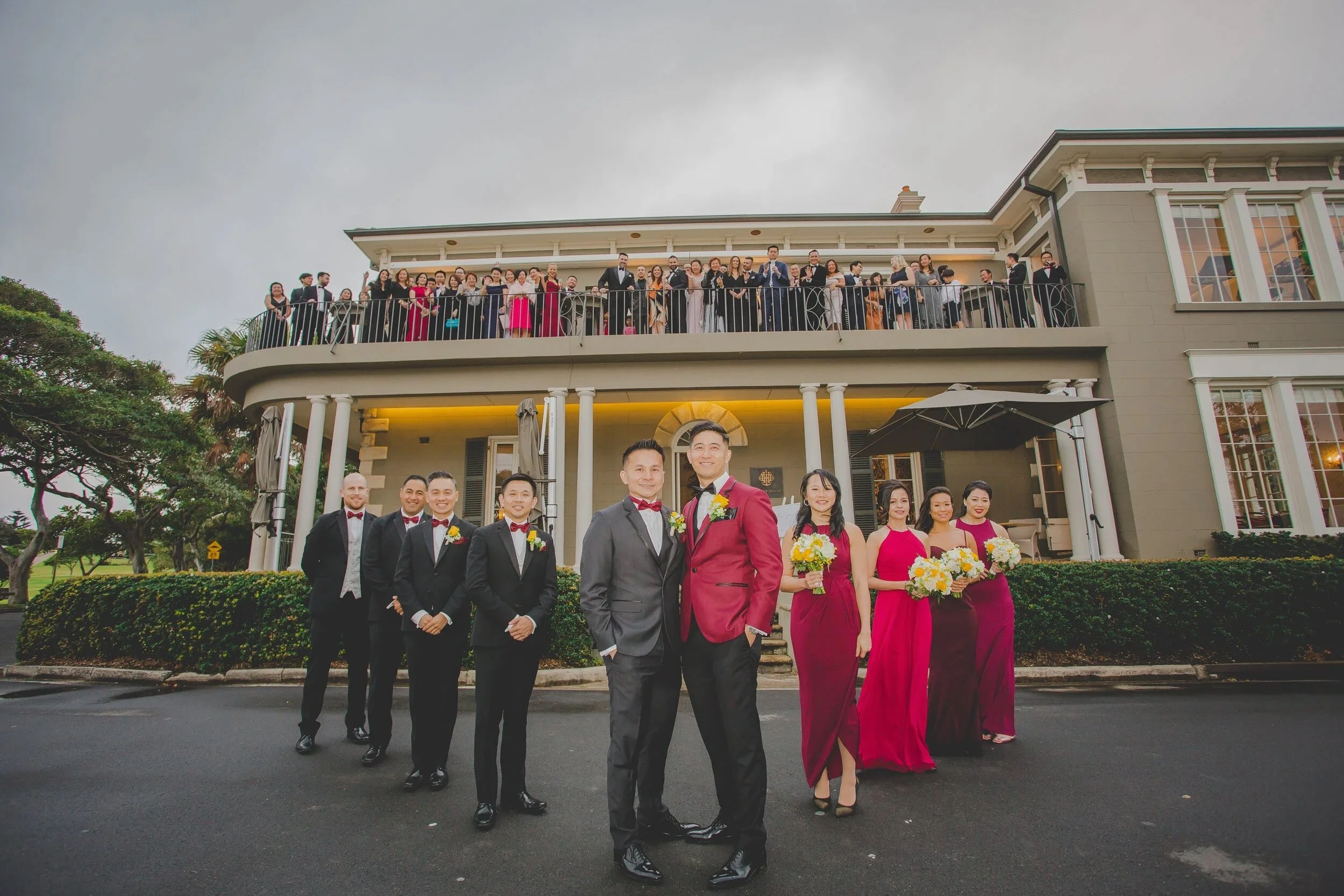 A large group of wedding guests gathered on and in front of a balcony of a large, historic style building during a wedding celebration. The wedding party, including the bride and groom, are standing in front, dressed in formal attire. The bride and g