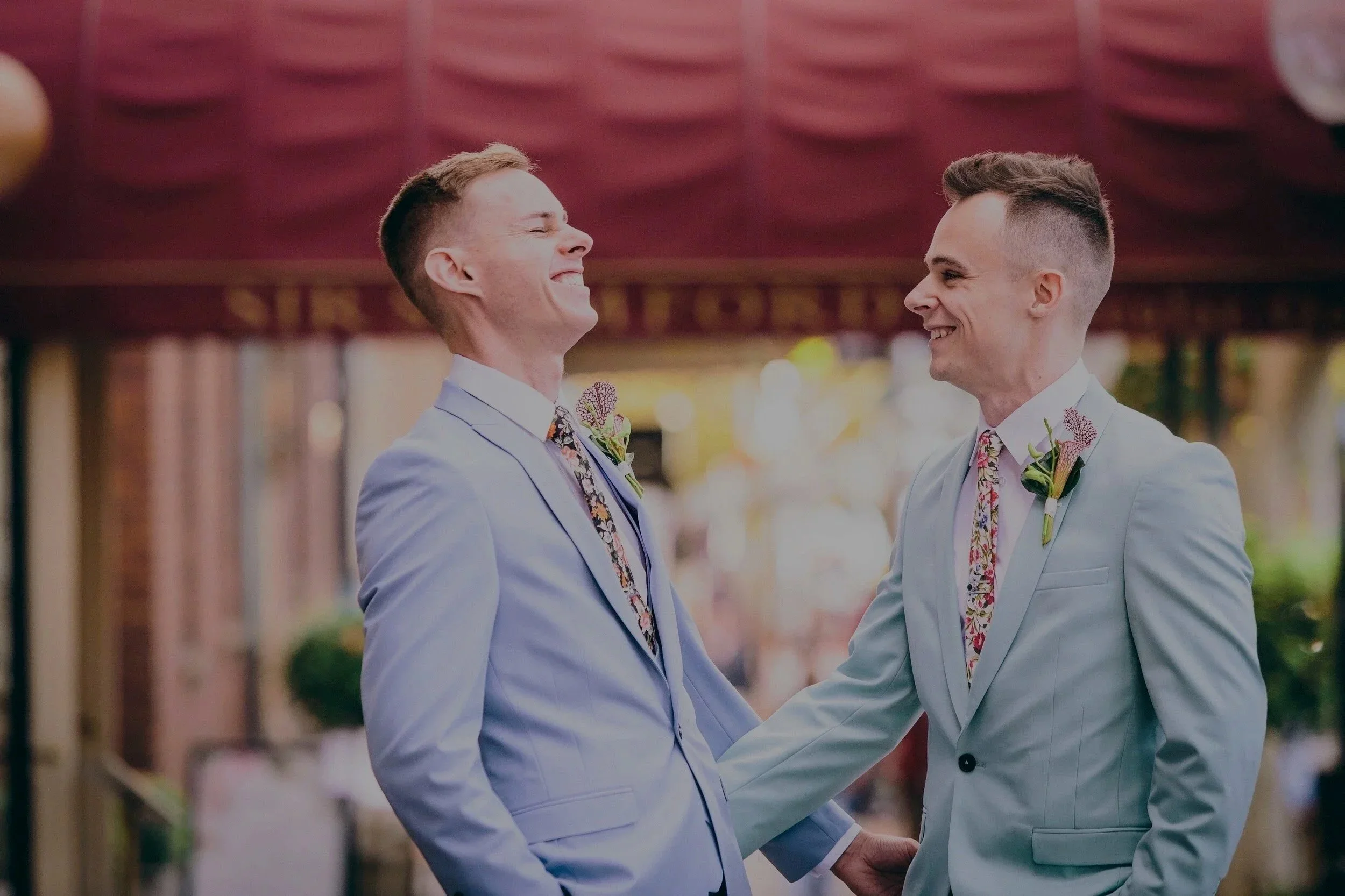 Two men in light suits smiling at each other at outdoor wedding, holding hands.