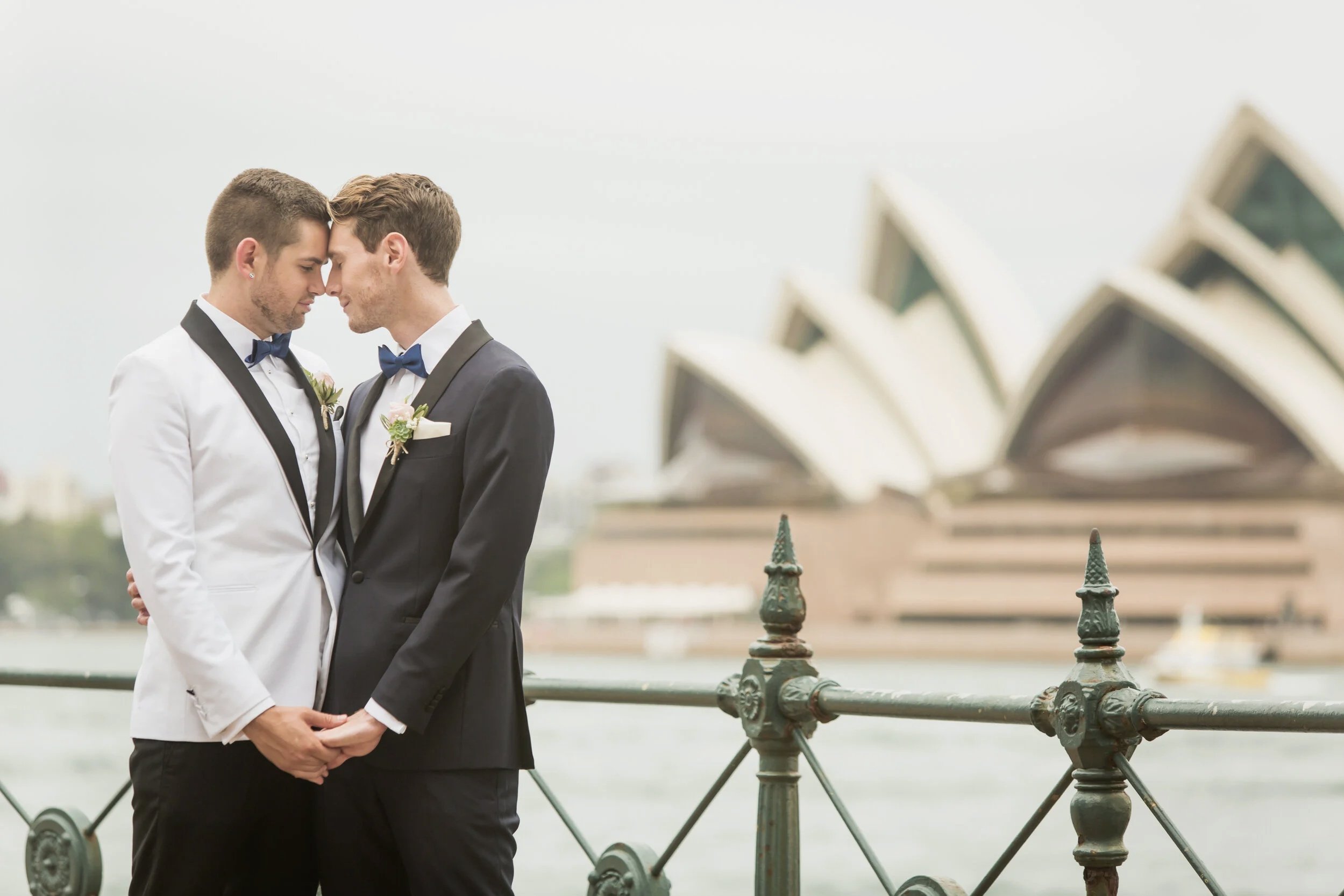 Two men dressed in tuxedos, one in white and the other in black, leaning foreheads together outdoors with the Sydney Opera House in the background, during a wedding or special event.