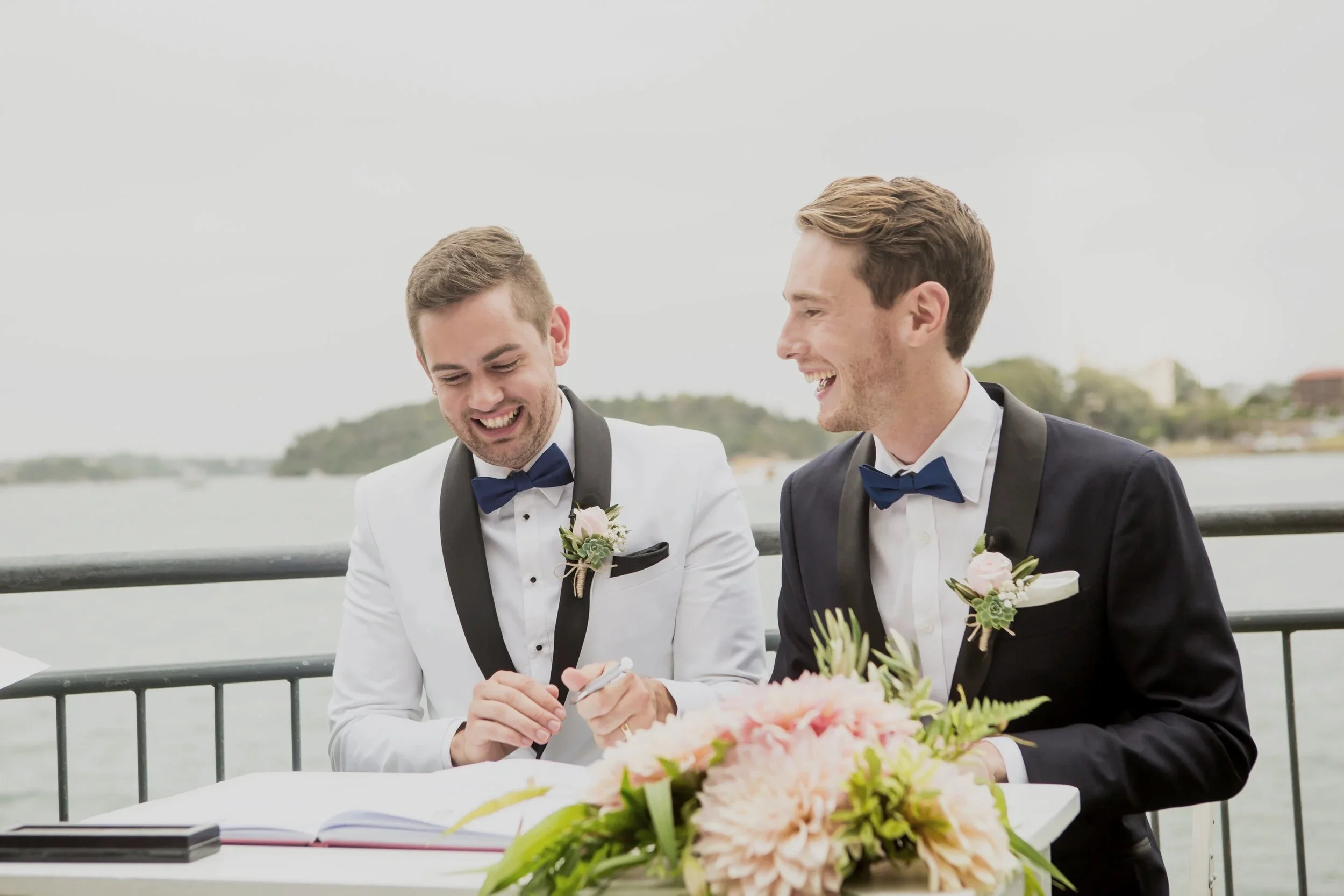 Two men dressed in tuxedos, one in white and one in black, smiling and signing a document at a table with a bouquet of pink and white flowers outdoors near water.