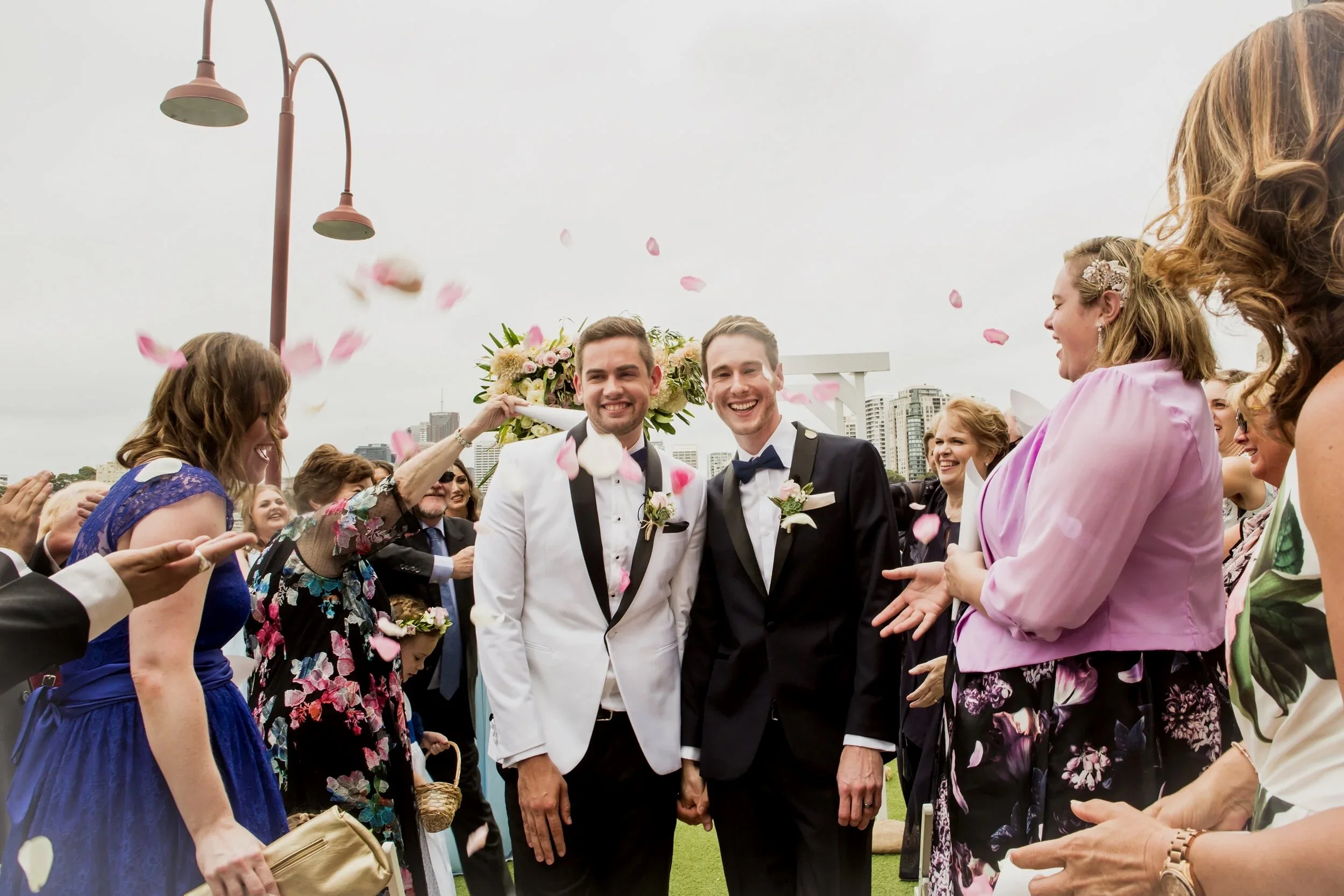 Two men in tuxedos smiling as they walk hand-in-hand through a shower of pink flower petals, surrounded by smiling women and onlookers at an outdoor wedding celebration, with city buildings in the background.