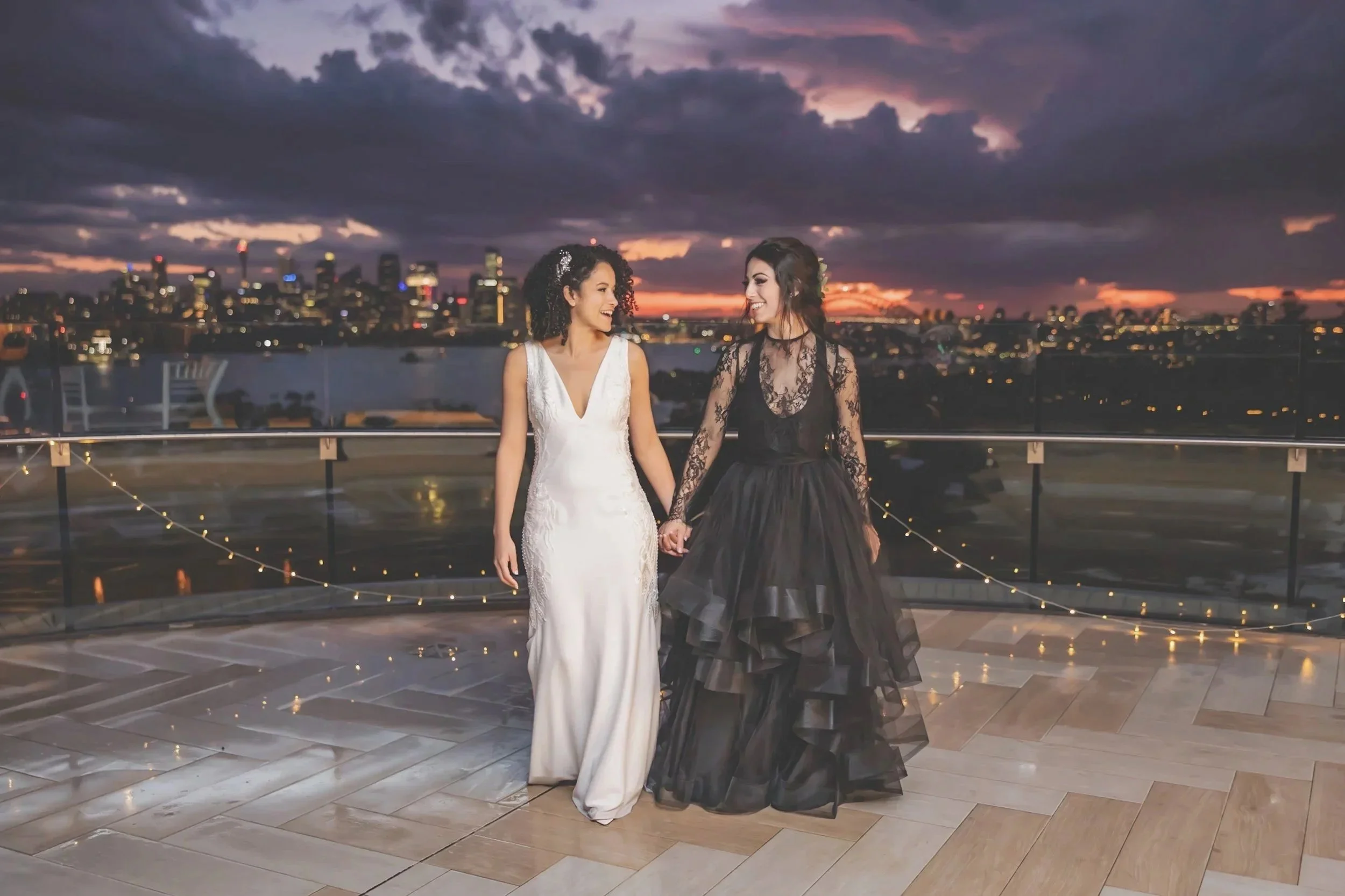 Two women in bridesmaid dresses holding hands on a rooftop at sunset, with city skyline in the background and string lights on the railing.