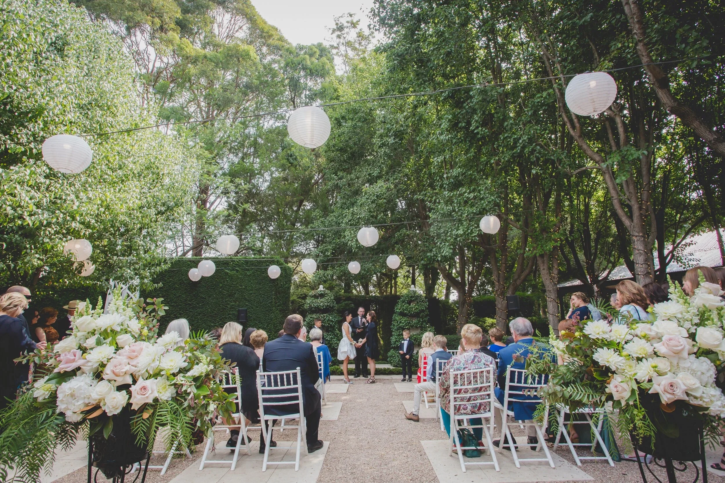 Outdoor wedding ceremony in a garden with white paper lanterns hanging from trees, guests sitting on white chairs, and a couple standing with an officiant at the altar, surrounded by lush greenery and floral arrangements.