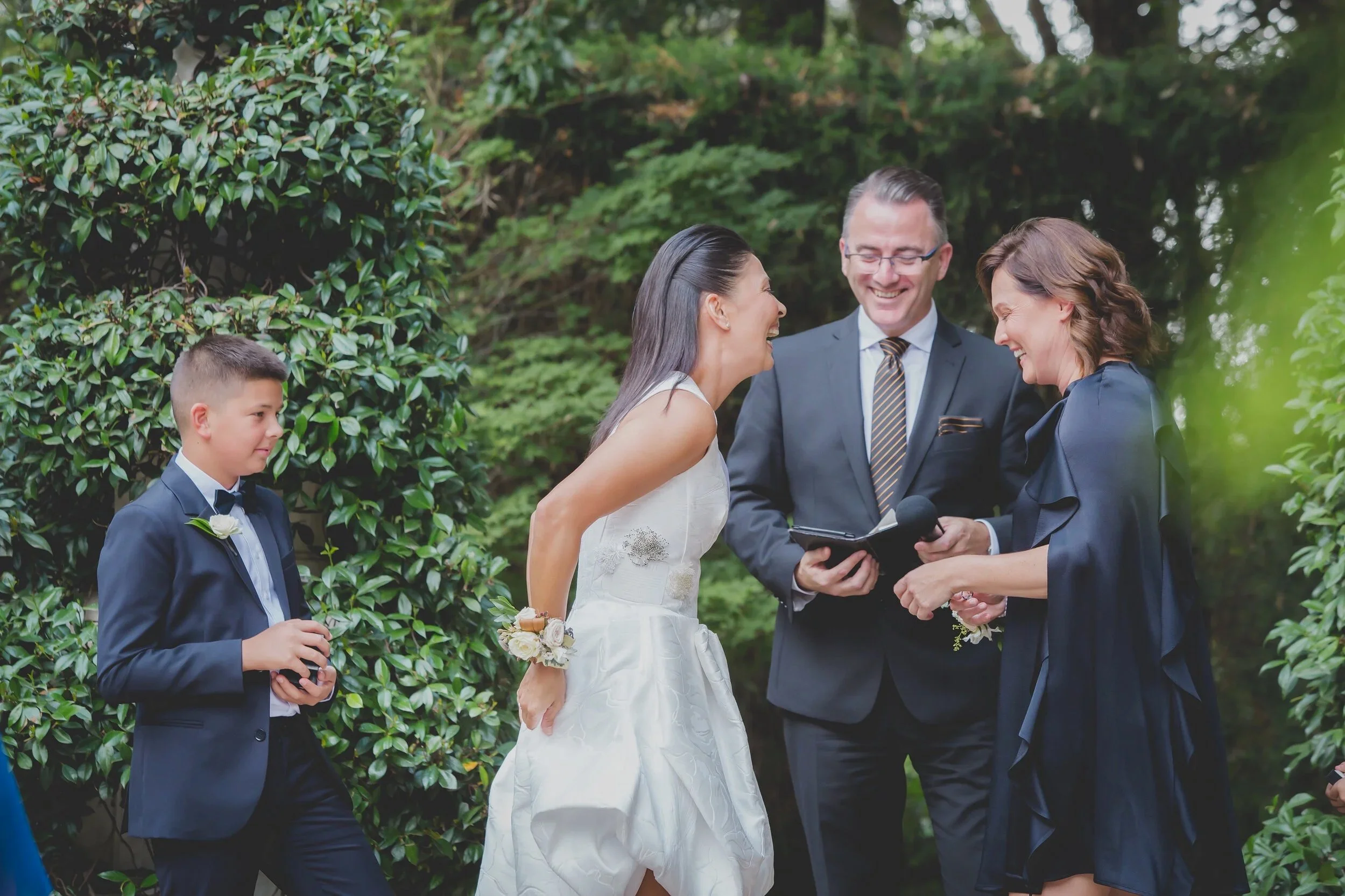 A wedding ceremony outdoors with the bride and groom smiling at each other, surrounded by greenery, during their vows or exchange of rings. A boy in a suit holds a ring box, and a woman in black appears to officiate the ceremony.