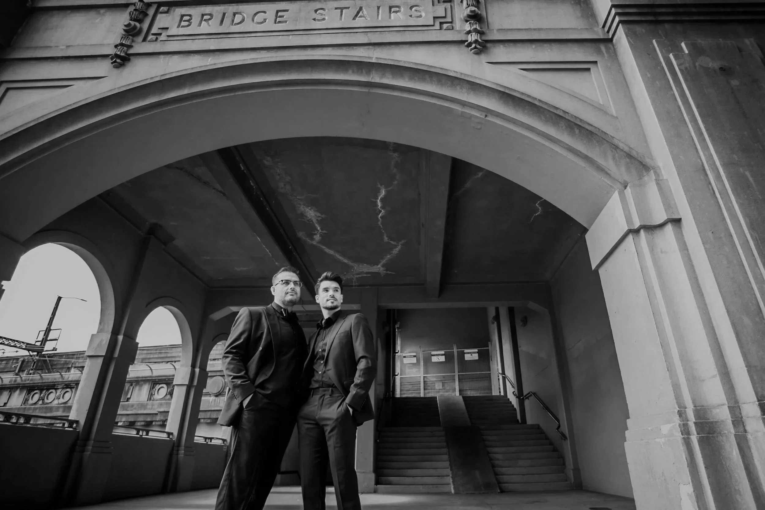 Two men in suits standing under the archway of a bridge with stairs leading up behind them.