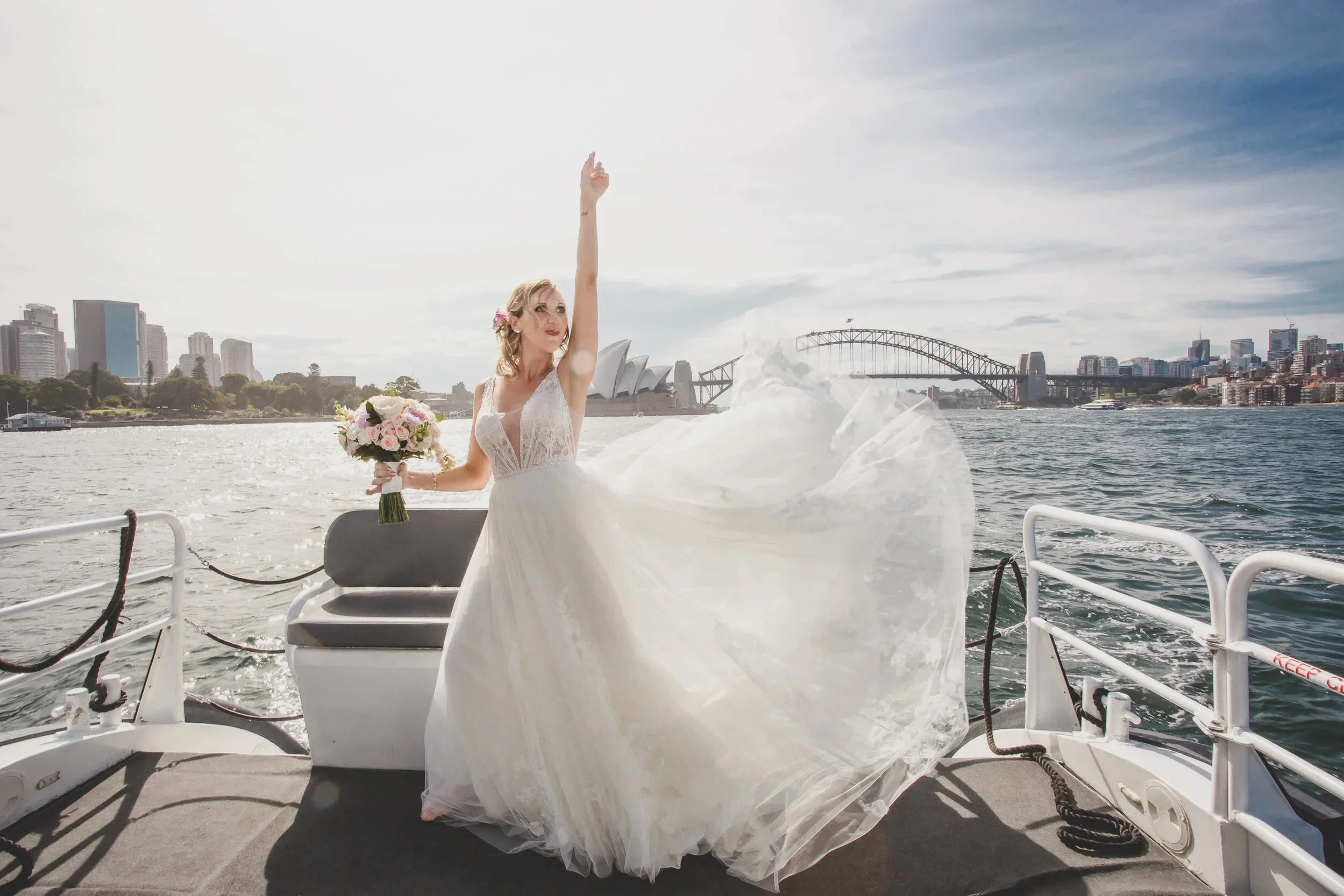 Bride on a boat with the Sydney Harbour Bridge and Opera House in the background, holding a bouquet of flowers and raising her right hand, with her wedding dress flowing in the wind.