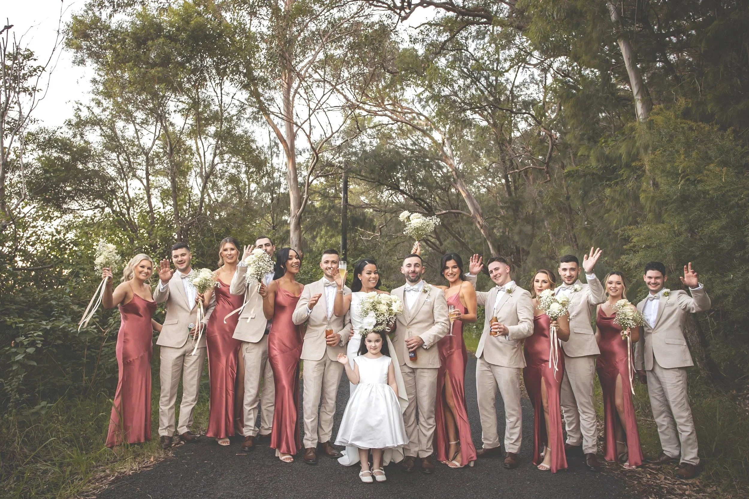 Wedding party of twelve people, including the bride in a white dress, groom in a beige suit, and ten bridesmaids and groomsmen, celebrating outdoors with trees in the background. Some are waving and holding wine bottles or bouquets.