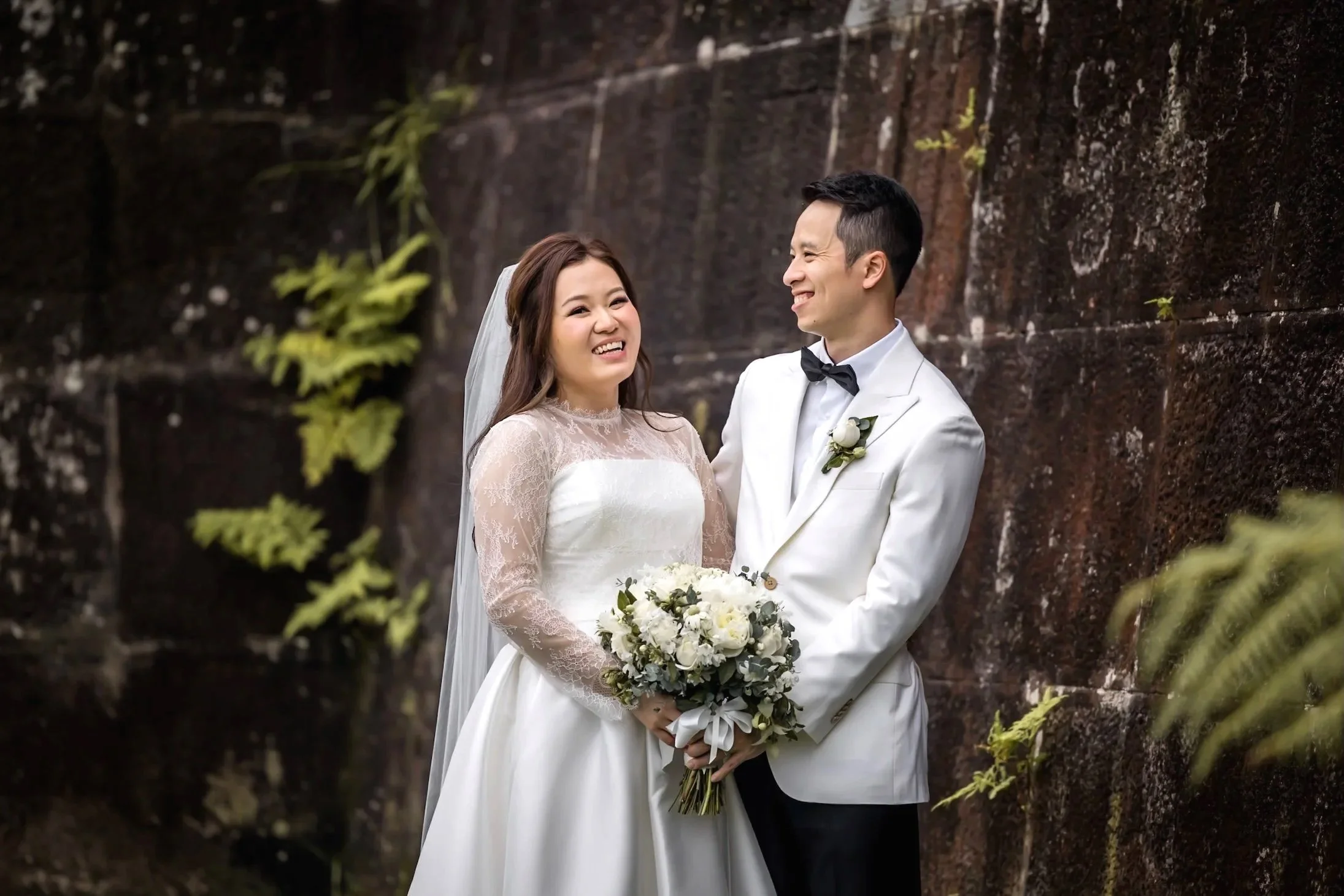 A bride and groom in wedding attire standing outdoors by a stone wall, smiling at each other. Gunners Barracks  Sydney - Katsu Nojiri Sydney Wedding Photographer
