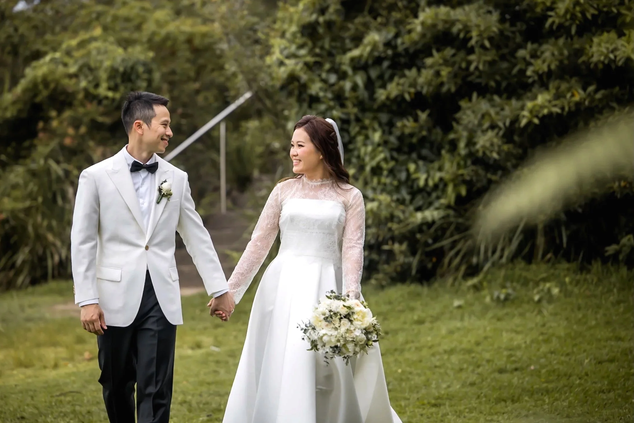 A bride and groom holding hands and smiling in a lush outdoor setting, the bride in a white lace wedding dress holding a bouquet of white flowers, Gunners Barracks  Sydney - Katsu Nojiri Sydney Wedding Photographer