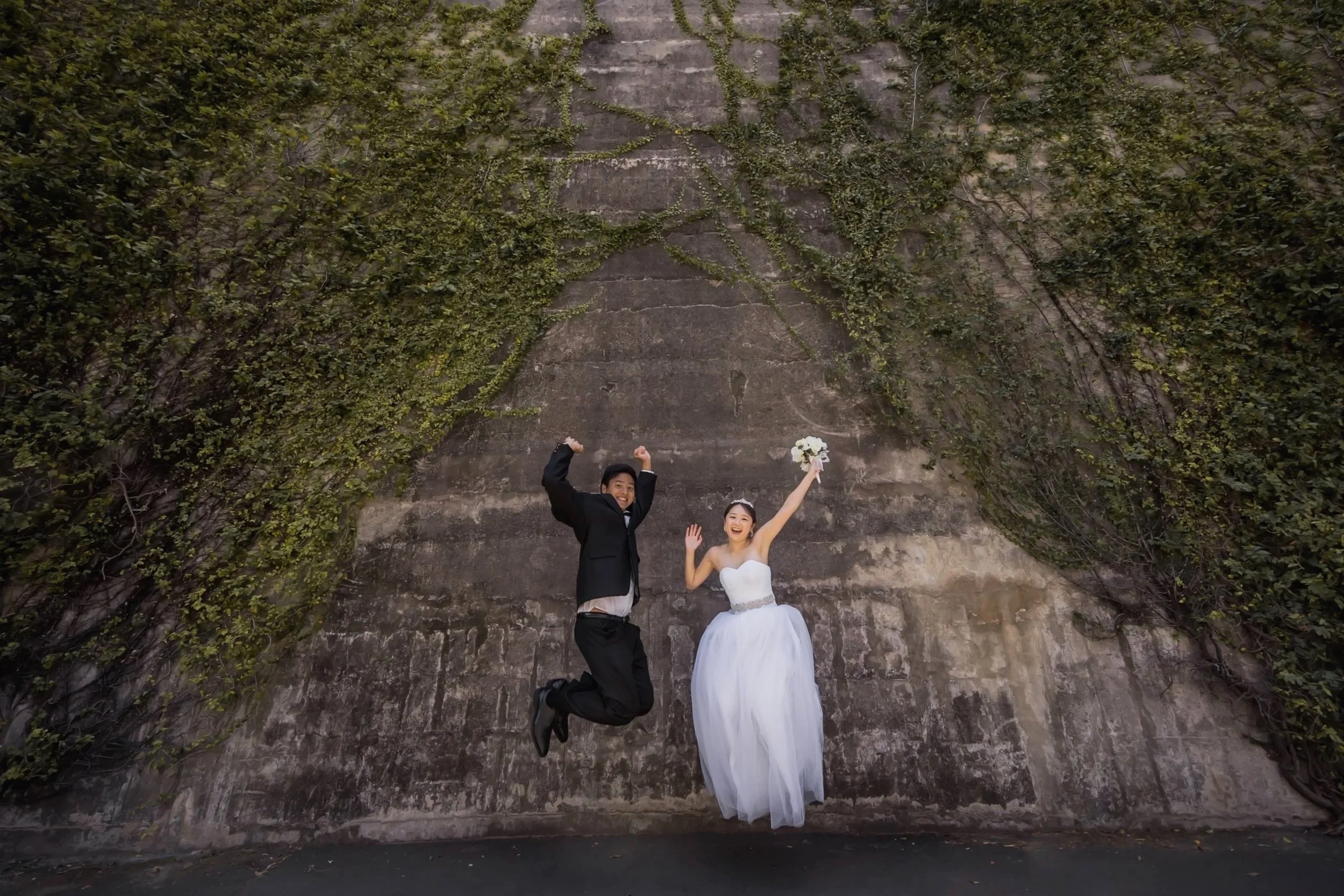 Bride and groom celebrating outdoors, bride holding a bouquet, jumping in the air, with a large ivy-covered wall in the background.