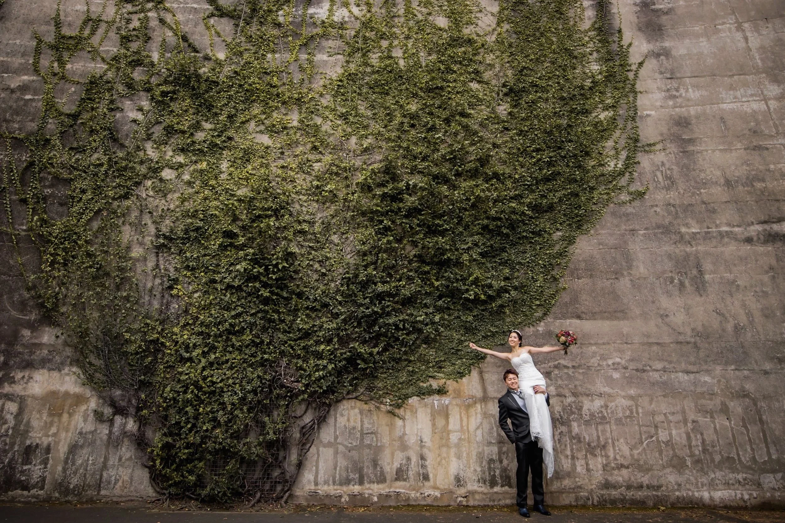 A bride in a white wedding dress is sitting on the shoulders of a groom in a black suit, standing against a large concrete wall overgrown with ivy. The bride is holding a bouquet of flowers and smiling with her arms outstretched.