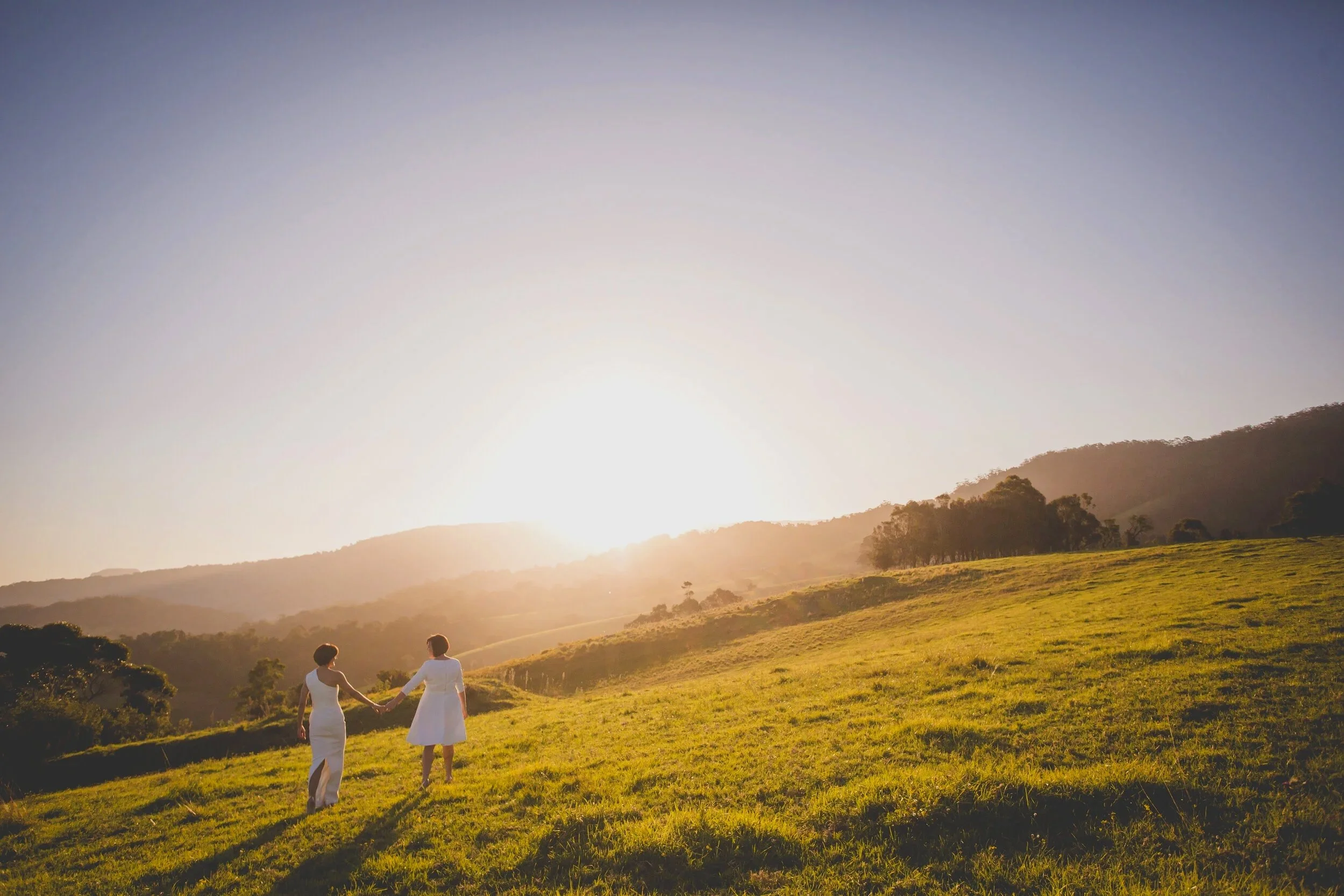 Two women in white dresses walking hand-in-hand through a grassy field during sunset.