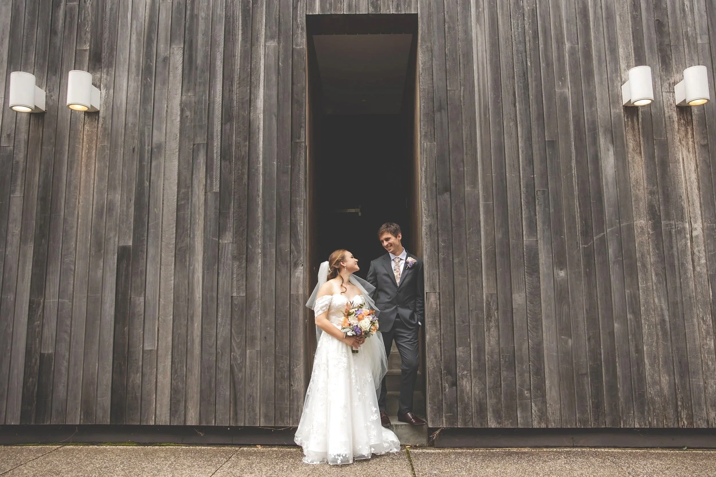 A bride and groom in wedding attire standing outside a large dark wooden building, sharing a happy moment.  - Sergeants Mess