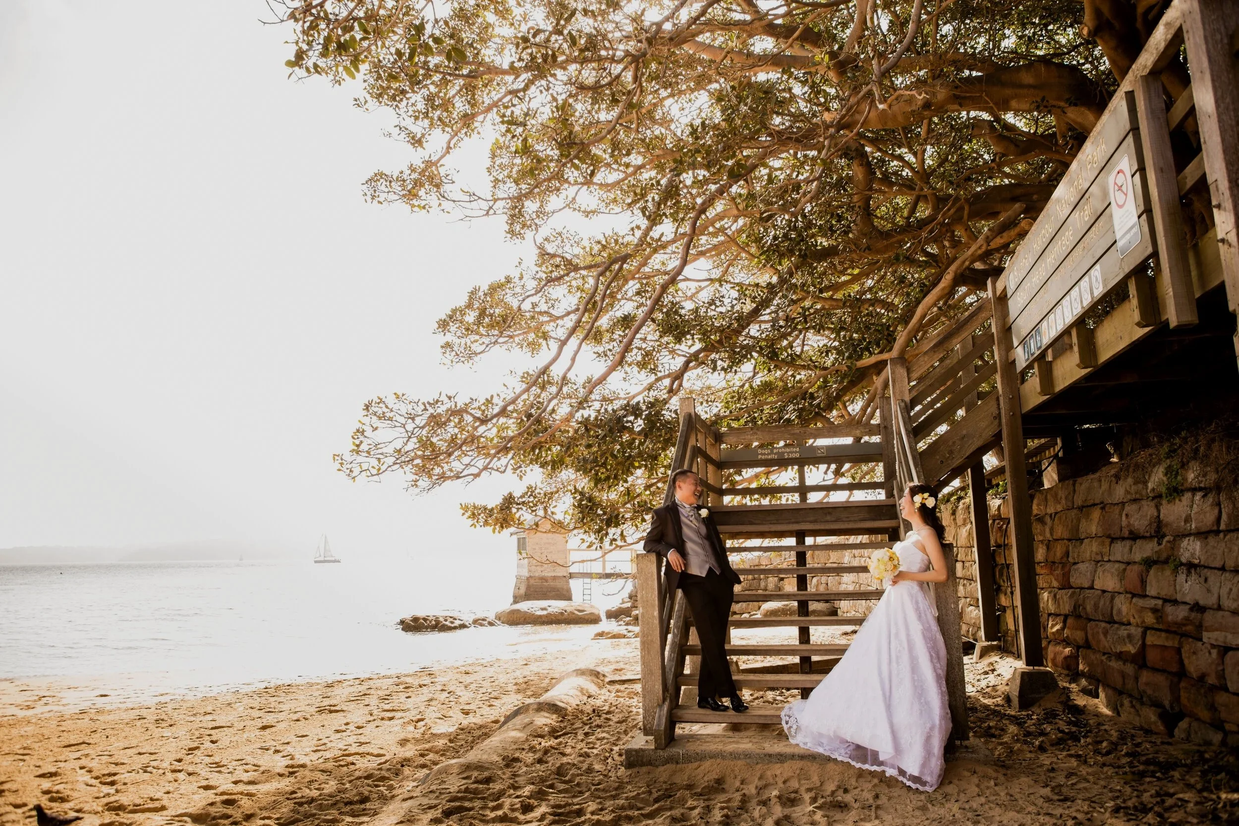 Bride and groom in wedding attire standing on a beach near a wooden staircase under a large tree, with the ocean and sailboats in the background.