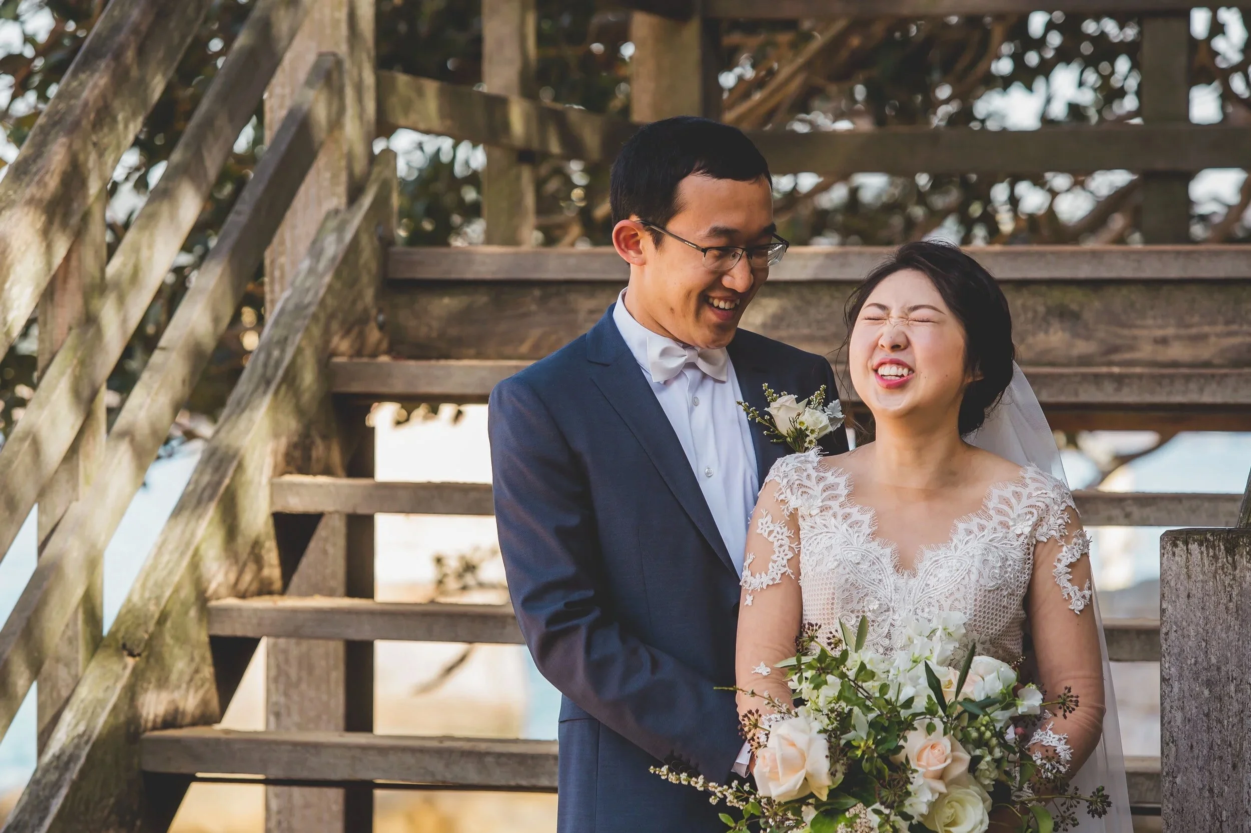 A bride and groom smiling happily during their wedding, standing on a wooden staircase with a detailed lace wedding dress and a dark suit, holding a bouquet of flowers.