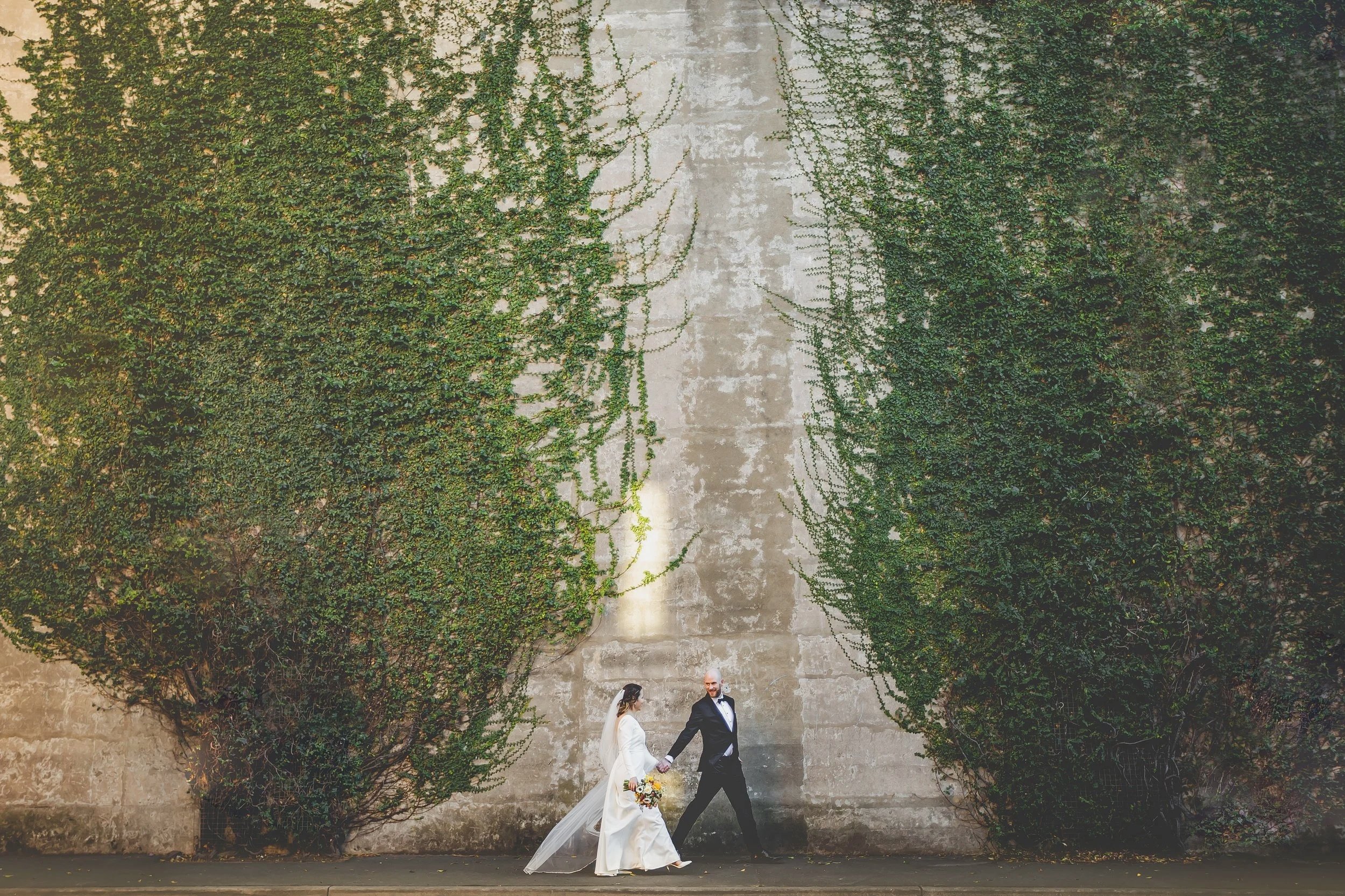A bride and groom walking hand in hand in front of a large concrete wall covered with green ivy, on a city street.