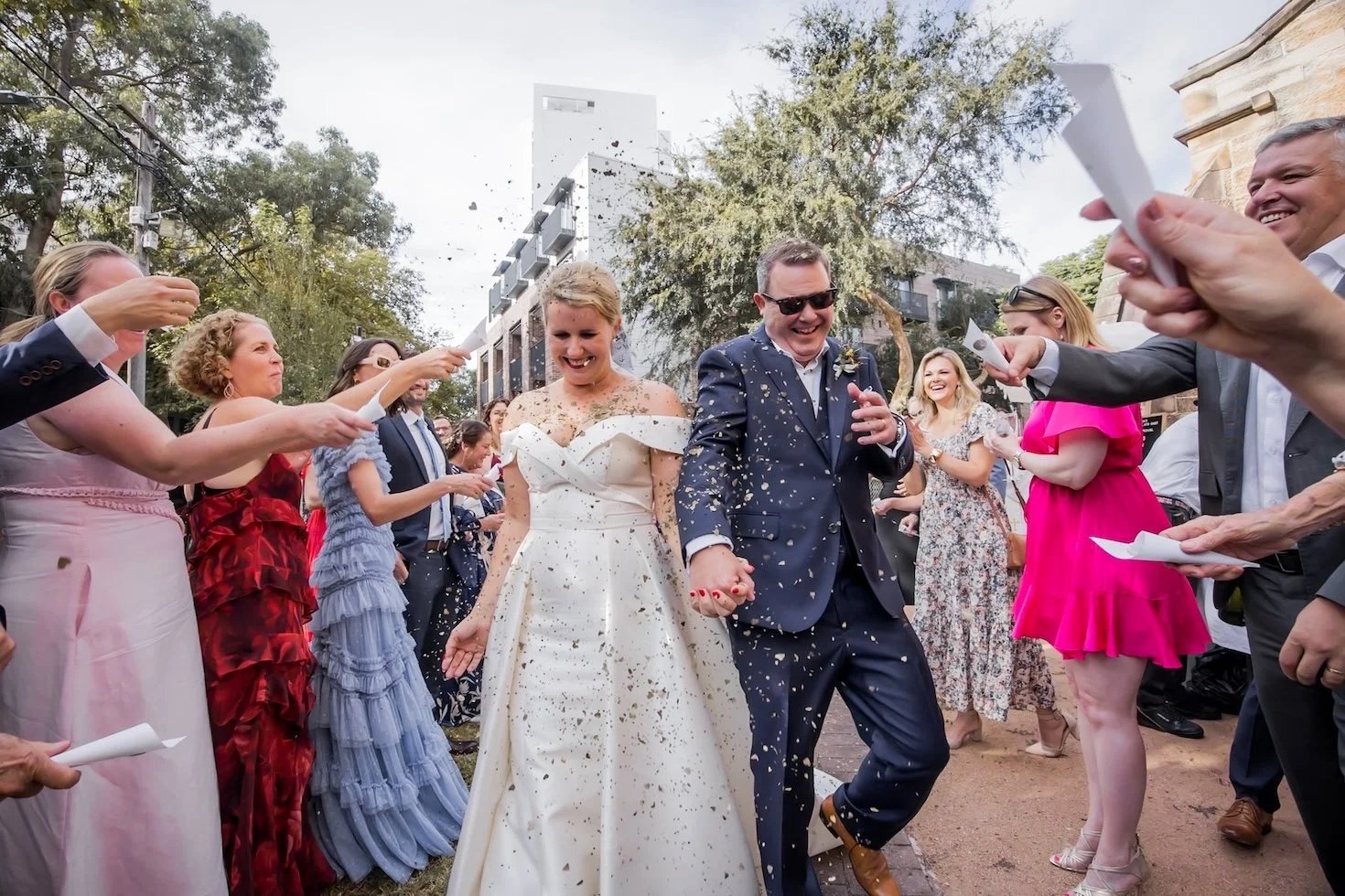 A newlywed couple in wedding attire walking through a crowd of guests at an outdoor celebration, with guests throwing confetti and some holding papers.