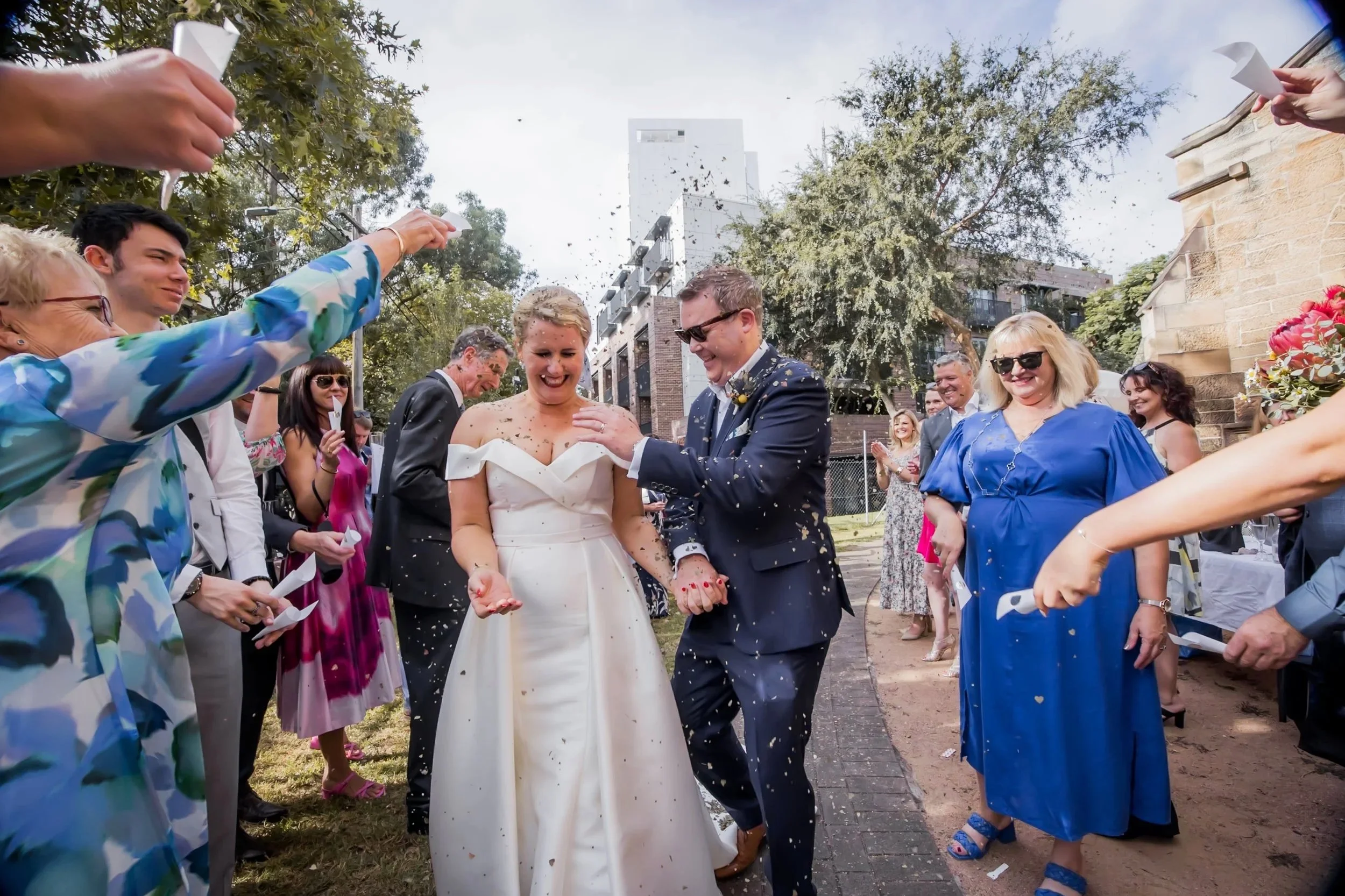 A bride and groom celebrating their wedding outdoors while guests throw confetti around them.
