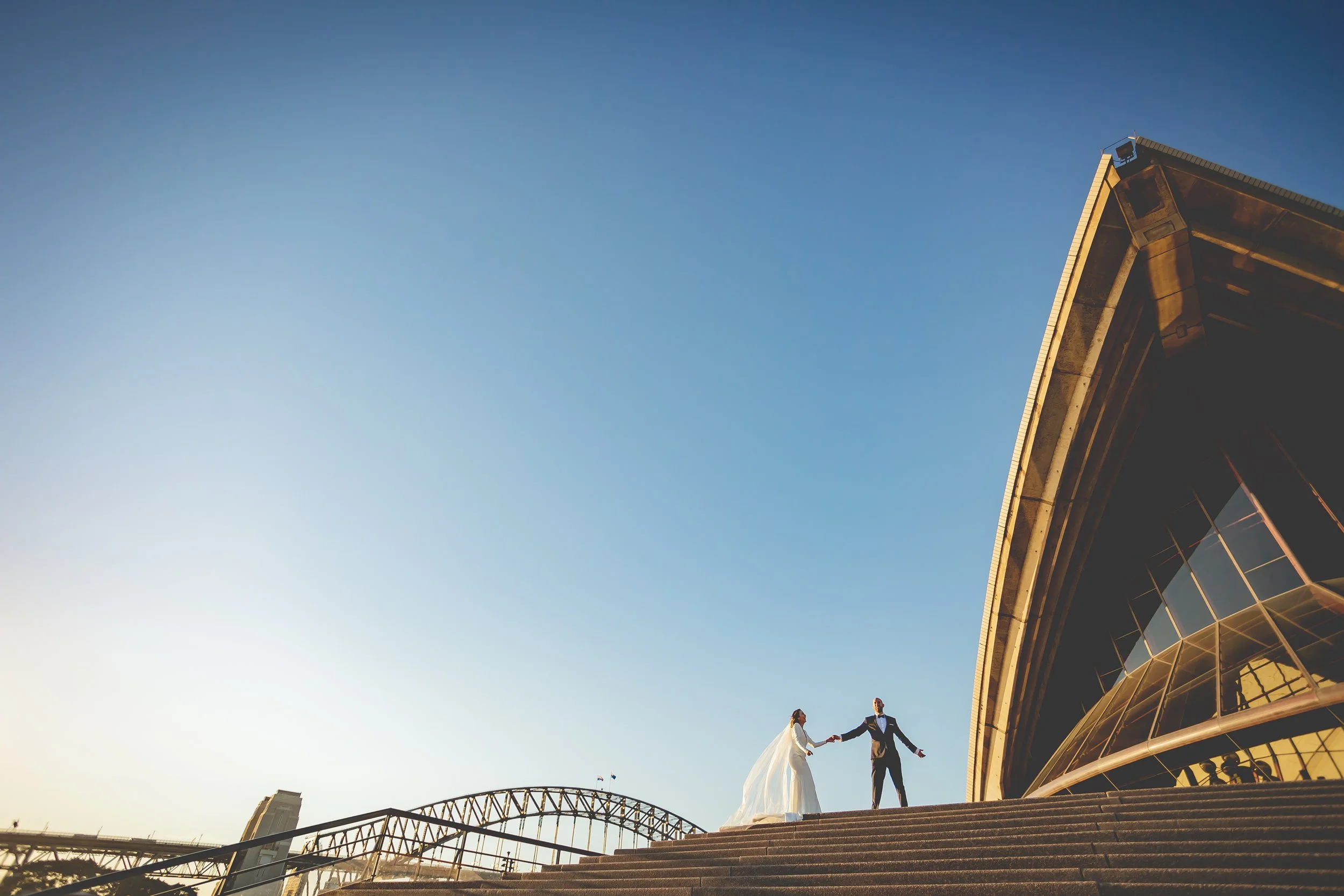 A bride and groom holding hands and dancing on a large staircase outside a modern building with large glass windows, with a bridge and city skyline in the background, during sunset. - Sydney Opera House