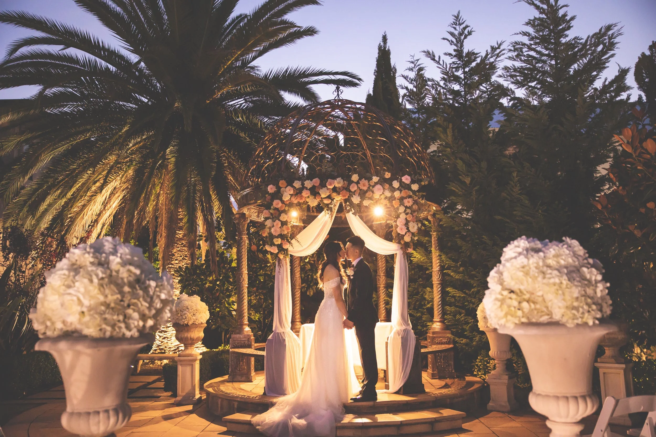 A bride and groom stand close together under an ornate gazebo decorated with flowers, illuminated by warm lights, during their wedding ceremony outdoors surrounded by palm trees and lush greenery at dusk.  Lauriston House Sydney - Katsu Nojiri Sydney