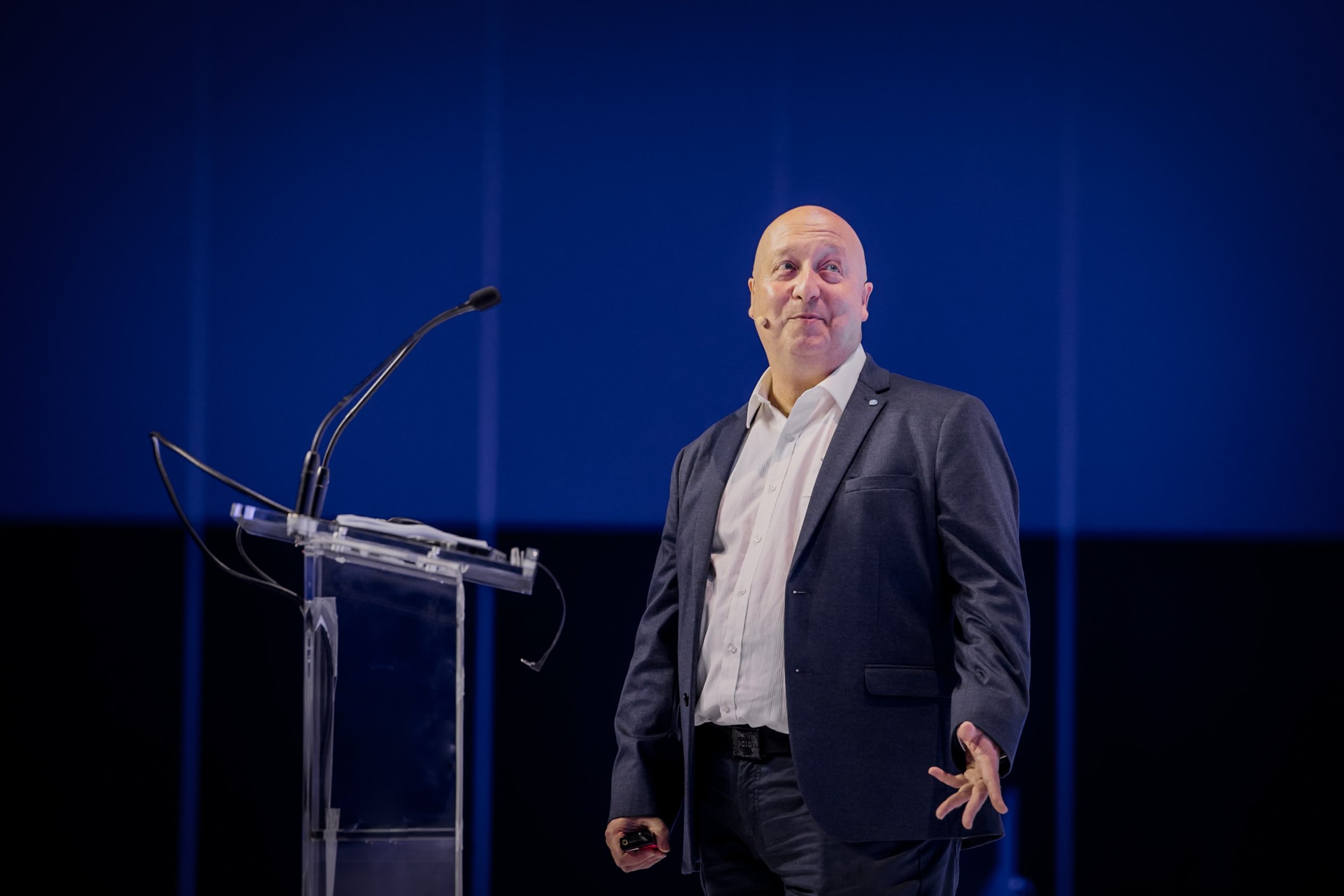 A bald man in a suit standing on a stage with a microphone and a clear podium, gesturing with his hand, in front of a blue background.