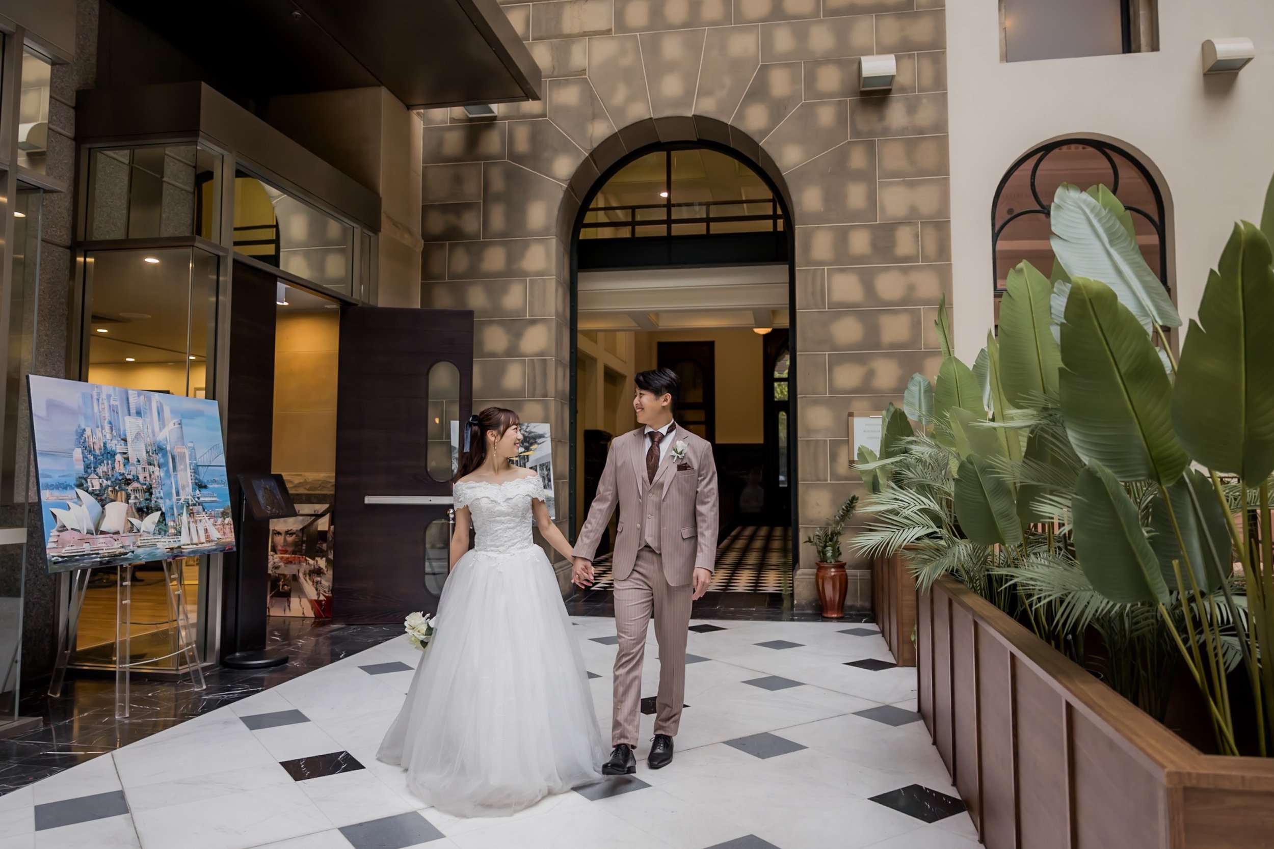 A bride and groom walking hand in hand in a hotel lobby or reception area. The bride is wearing a white wedding gown, and the groom is dressed in a light-colored suit with a tie. They are smiling at each other.