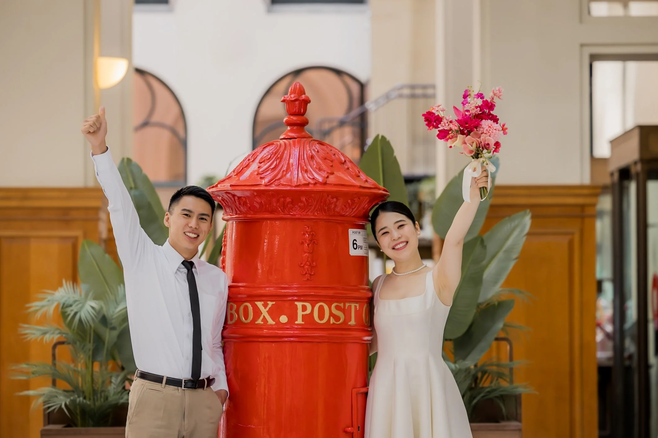 A happy couple standing next to a large red mailbox inside a building. The woman is holding a bouquet of pink flowers, and the man has his hand raised in celebration. Both are smiling.