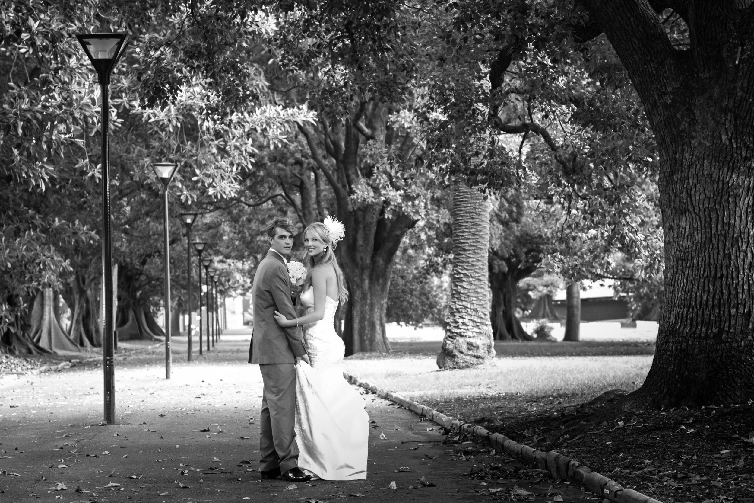 A black and white photograph of a newly married couple holding hands and smiling in a park lined with trees and street lamps.