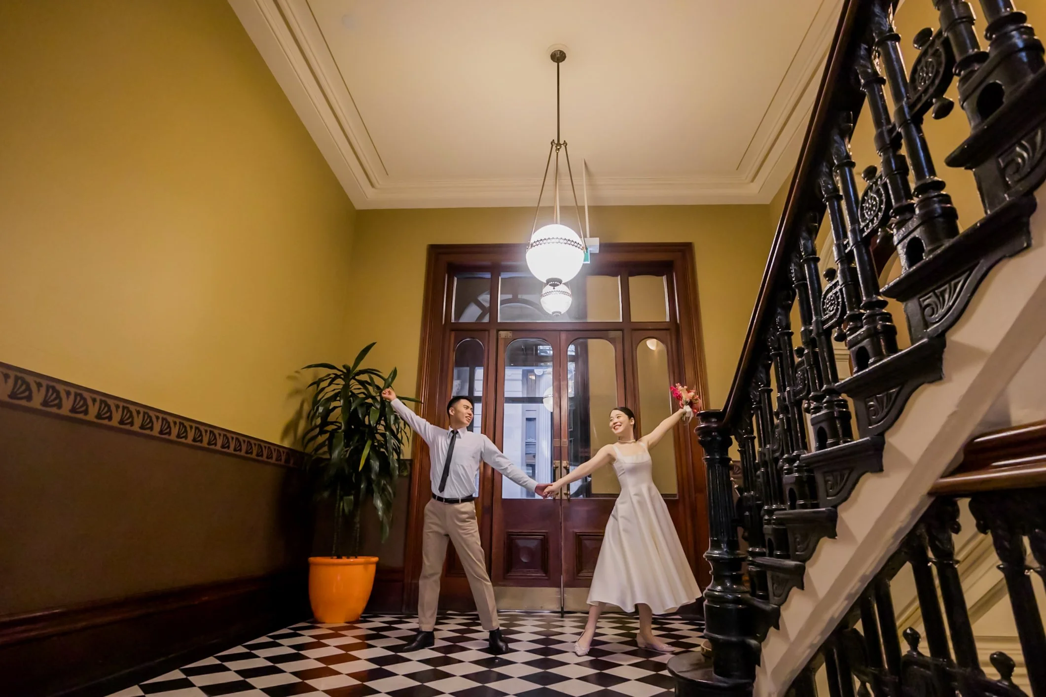 A couple dancing inside a warmly lit, vintage-style building with high ceilings, a large wooden door, a black ornate staircase, and a tall potted plant.