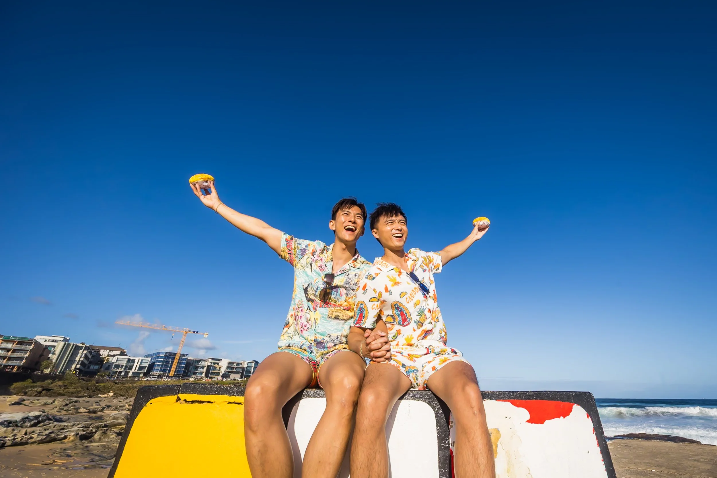 Two young men in colorful Hawaiian shirts sitting on a barrier at the beach, holding donuts, smiling, with the ocean and a clear blue sky in the background. Maroubra Beach  Sydney - Katsu Nojiri Sydney Wedding Photographer
