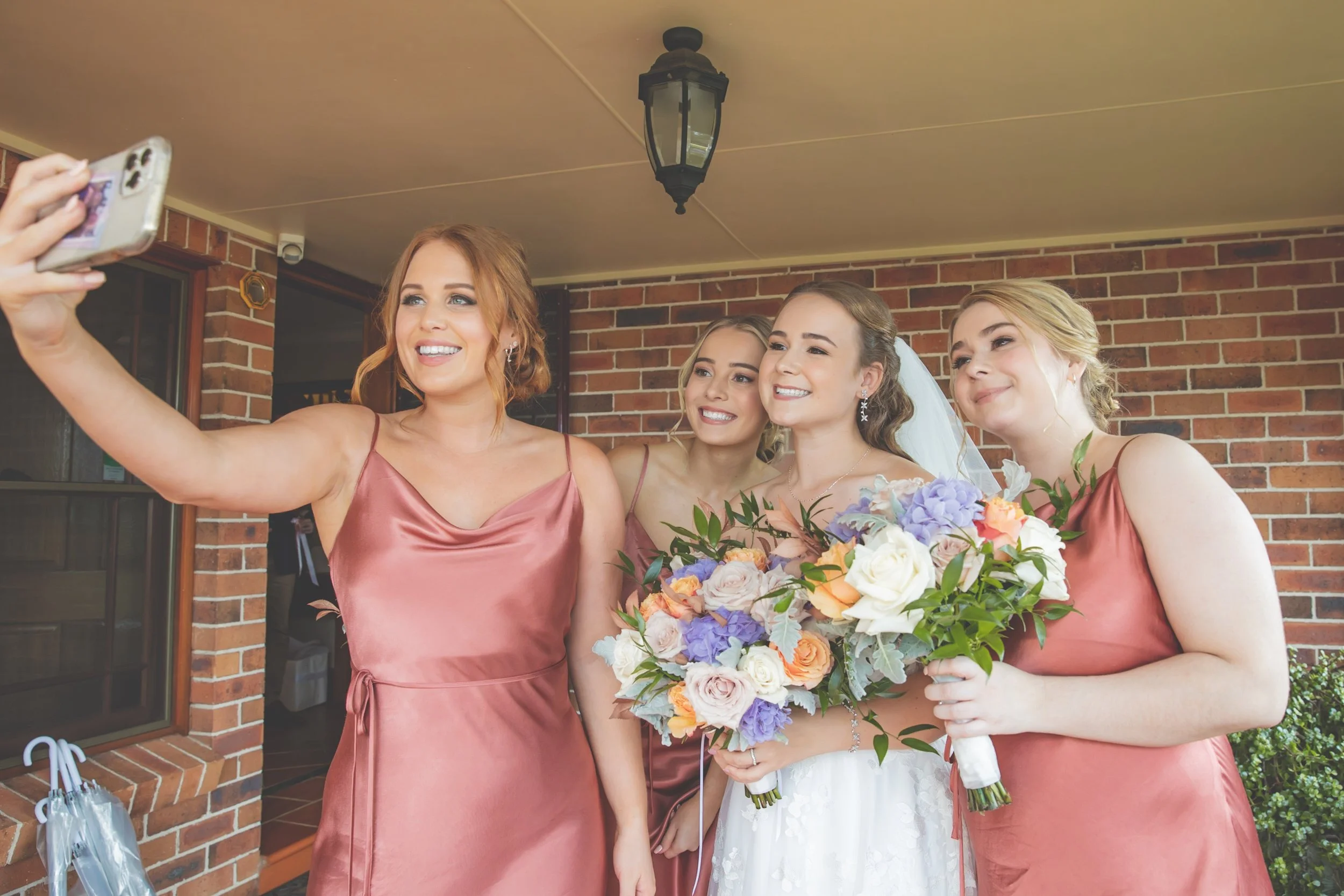 Group of four women, including a bride and bridesmaids, taking a selfie on the porch of a house with brick walls; the bride is holding a bouquet of flowers, all women are smiling.