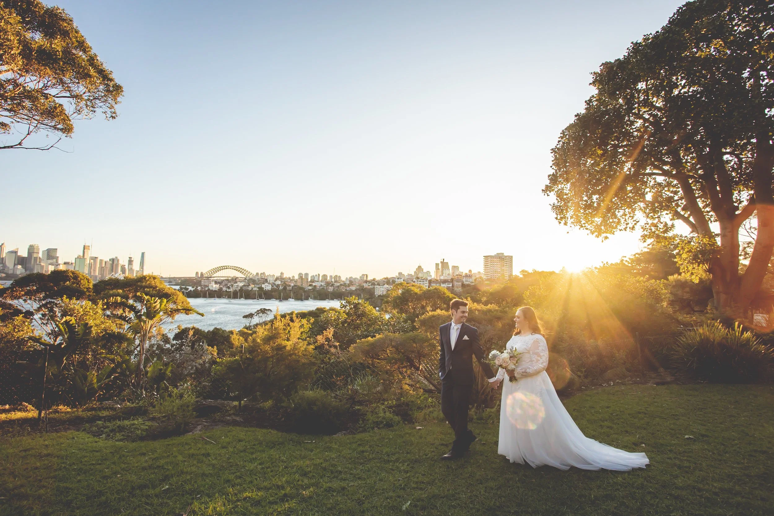 A bride and groom walking hand in hand outdoors at sunset with a city skyline and river in the background. - Taronga House Sydney - Katsu Nojiri Sydney Wedding Photographer