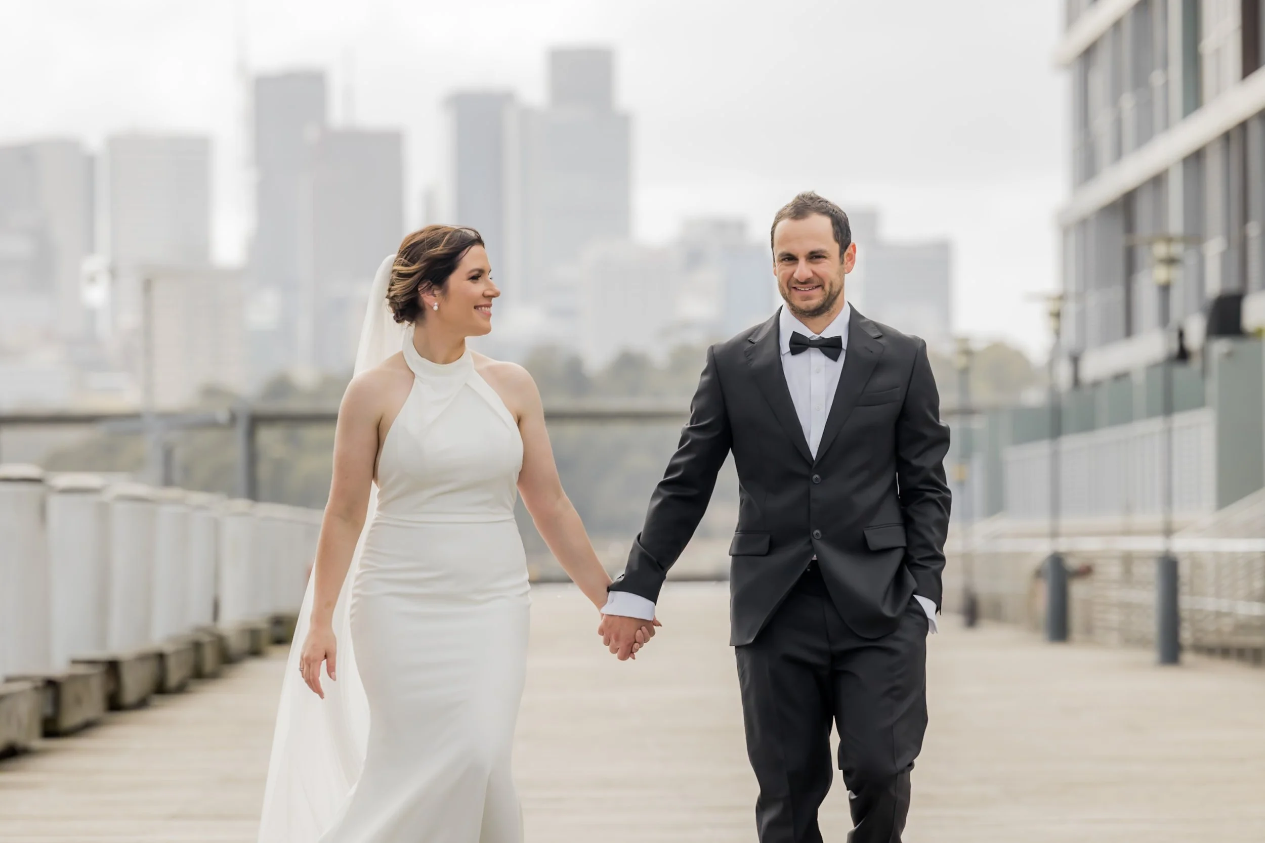 A bride and groom walking hand in hand outdoors on a city pier, dressed in wedding attire with city skyline in the background, smiling and looking joyful.