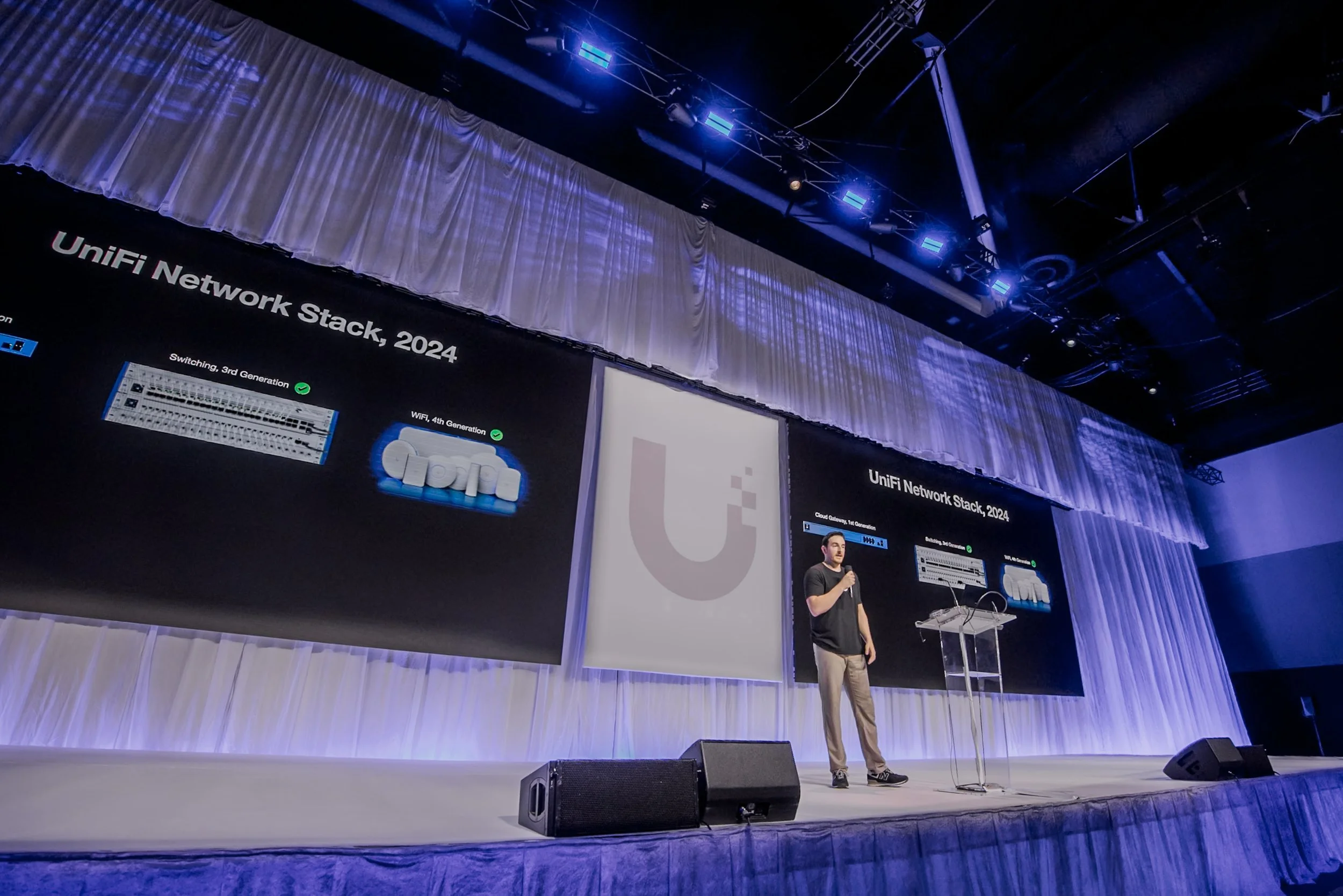 A person standing on a stage giving a presentation about the 2024 UniFi Network Stack, with large screens displaying technical diagrams and information behind him, illuminated by stage lighting.