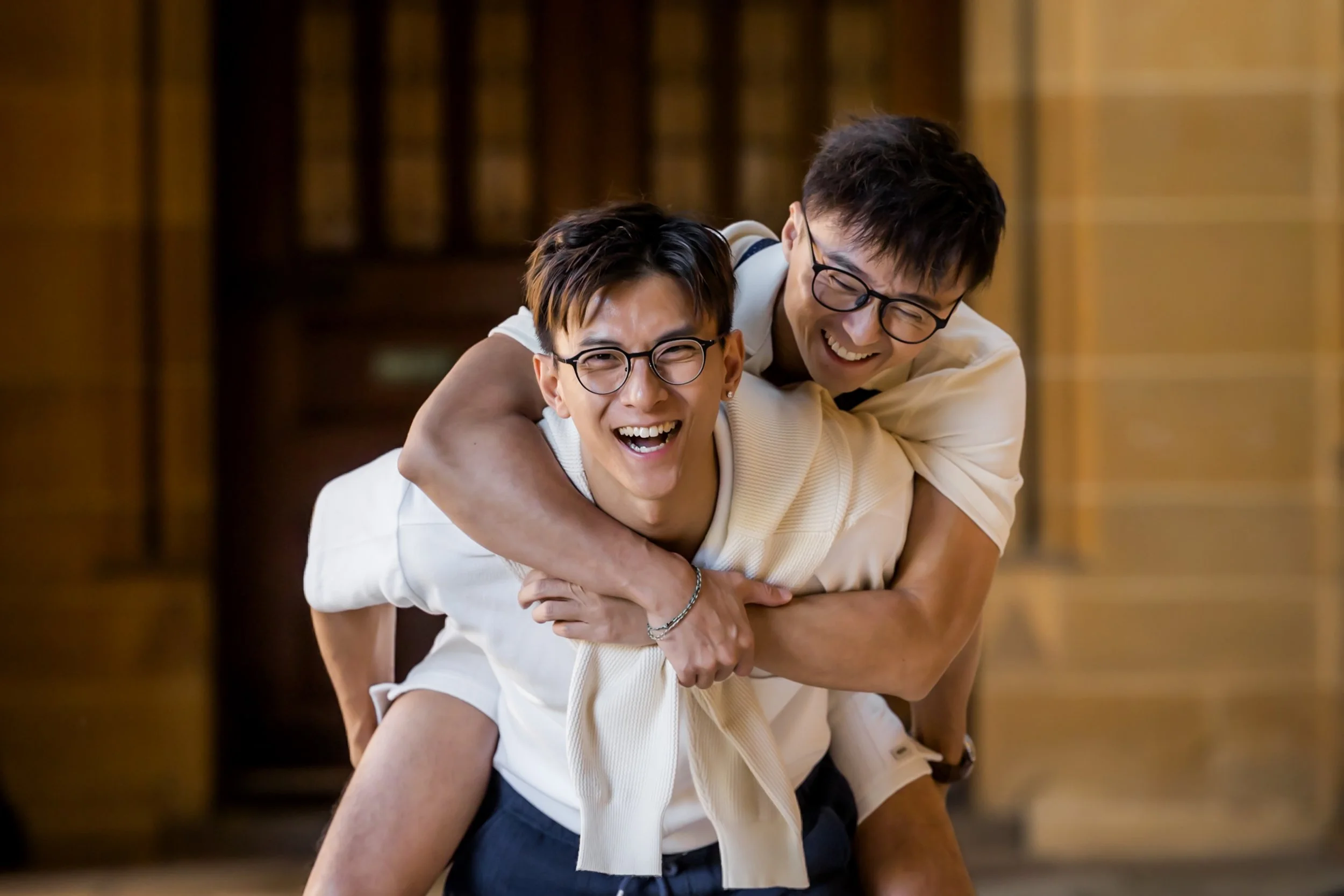 Two young men, both wearing glasses, happily playing and embracing each other indoors, smiling joyfully.