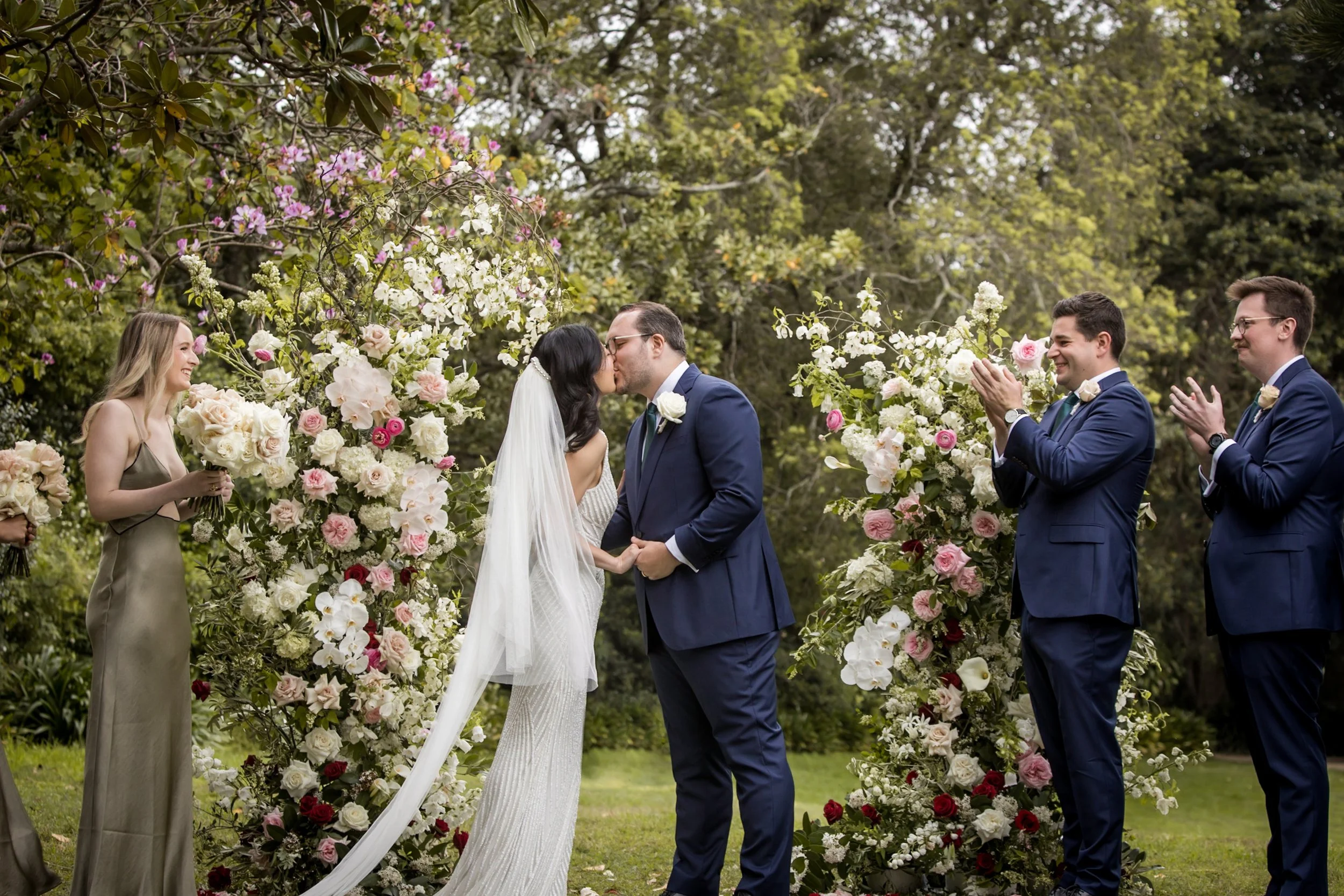 A bride and groom kissing during their outdoor wedding ceremony, surrounded by floral arrangements and a wooded background, with bridesmaids and groomsmen clapping. -Vaucluse House  Sydney - Katsu Nojiri Photographer