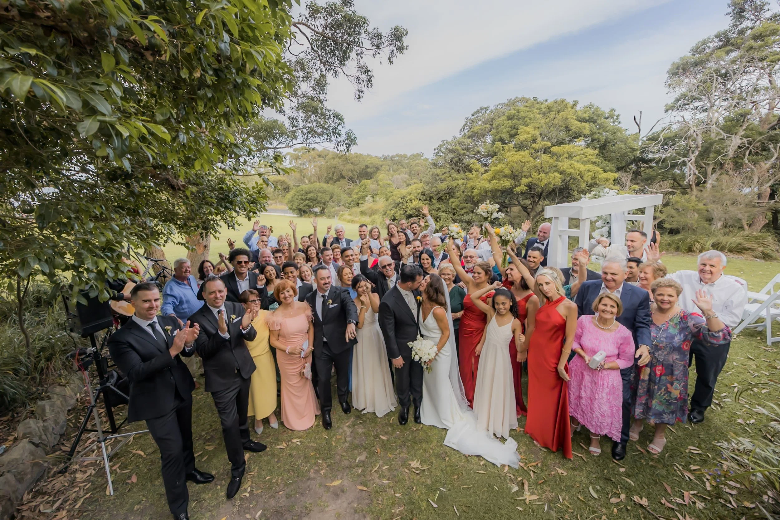 A large group of people gathered outdoors for a celebration, with a couple in wedding attire in the center surrounded by friends and family, under a blue sky and green trees.
