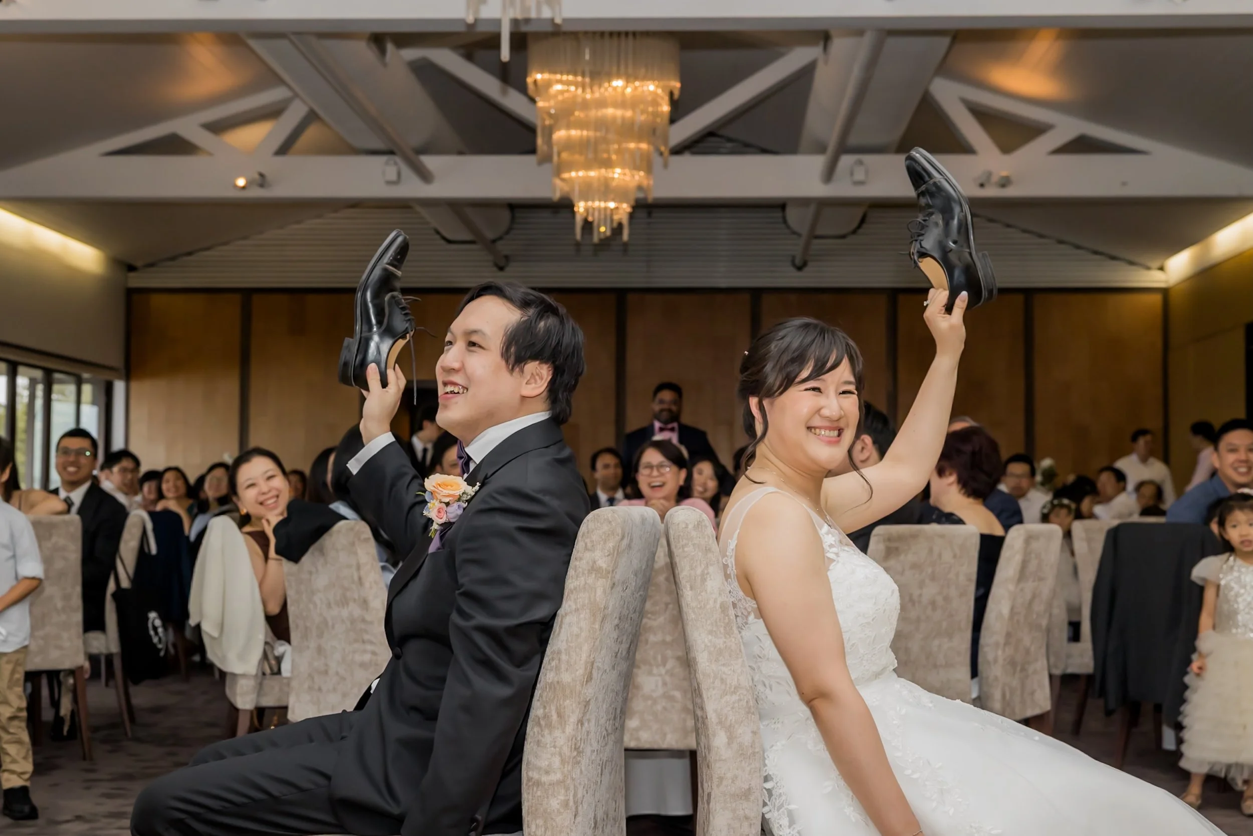 A bride and groom are playing a game at their wedding reception, sitting back to back and holding up shoes, smiling and laughing.